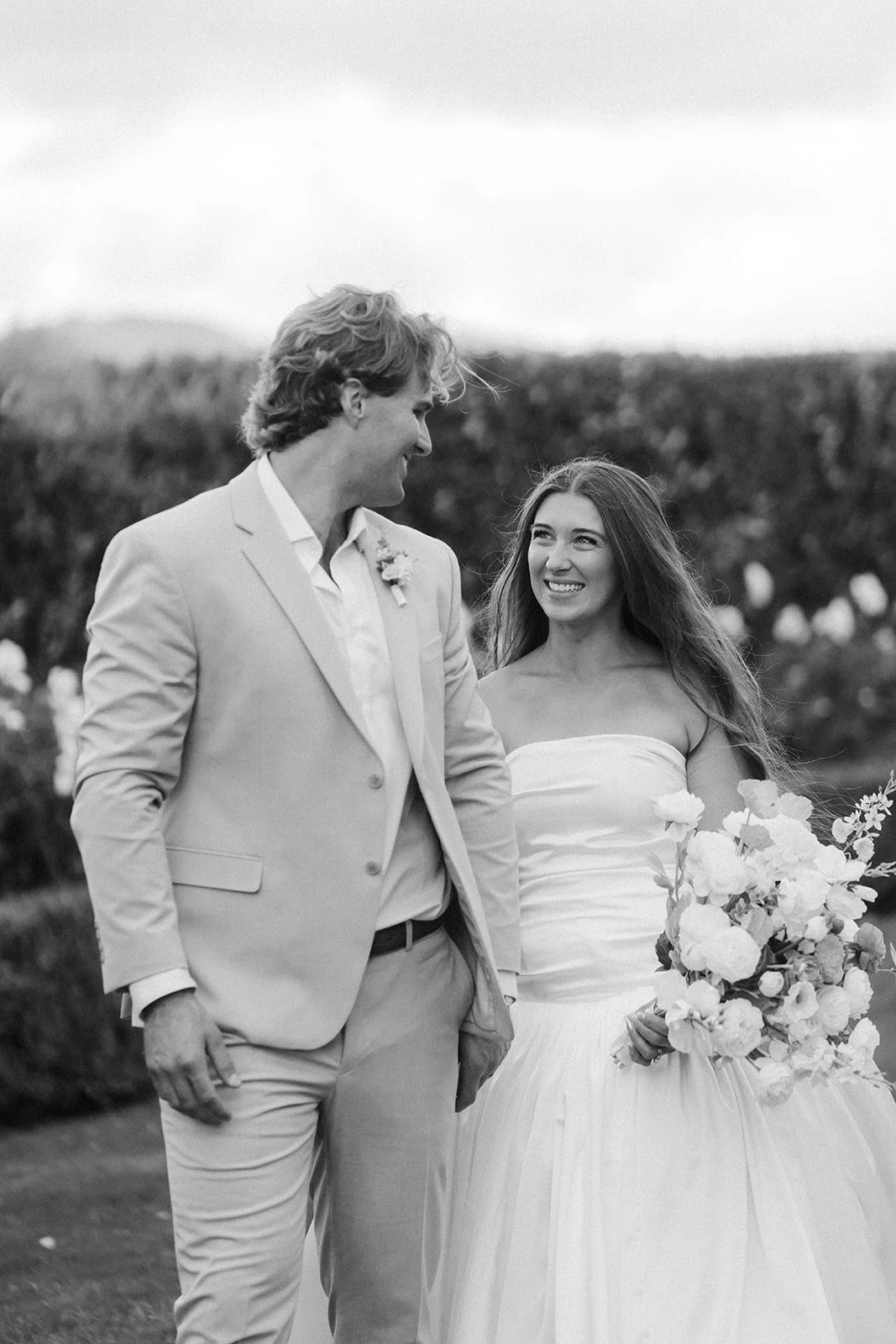 A black-and-white photograph of a smiling bride in a strapless wedding gown holding a bouquet of flowers, standing next to a groom in a light-colored suit, outdoors in a garden setting at San Marcos wedding venue.