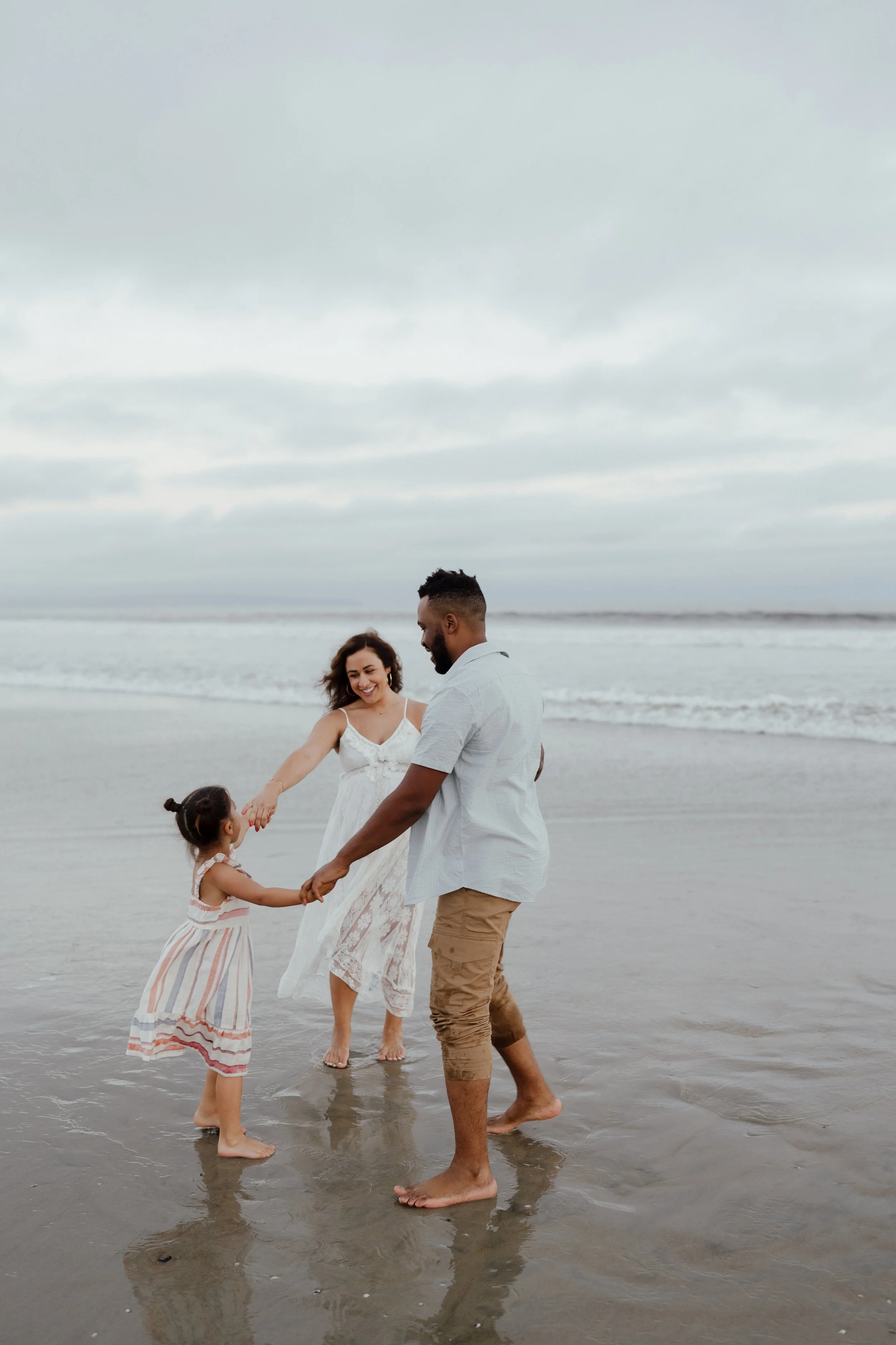 A family of three on the beach, holding hands and playing in the water, under a cloudy sky.