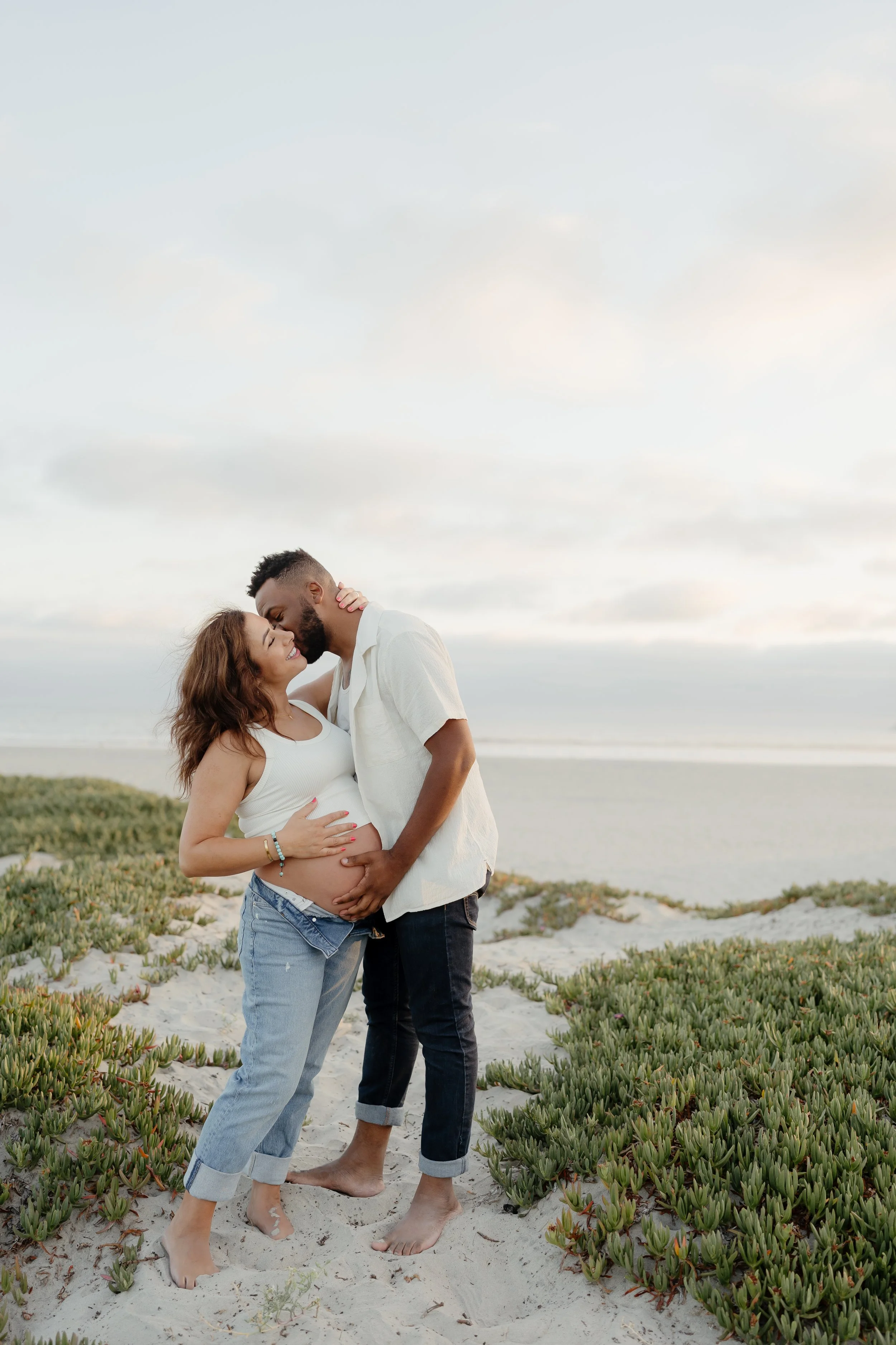 A pregnant woman and a man on a beach, touching foreheads and smiling. The woman has long brown hair and is wearing a white top and jeans. The man has a beard, short hair, and is wearing a white shirt and jeans. They are standing on sand near green p
