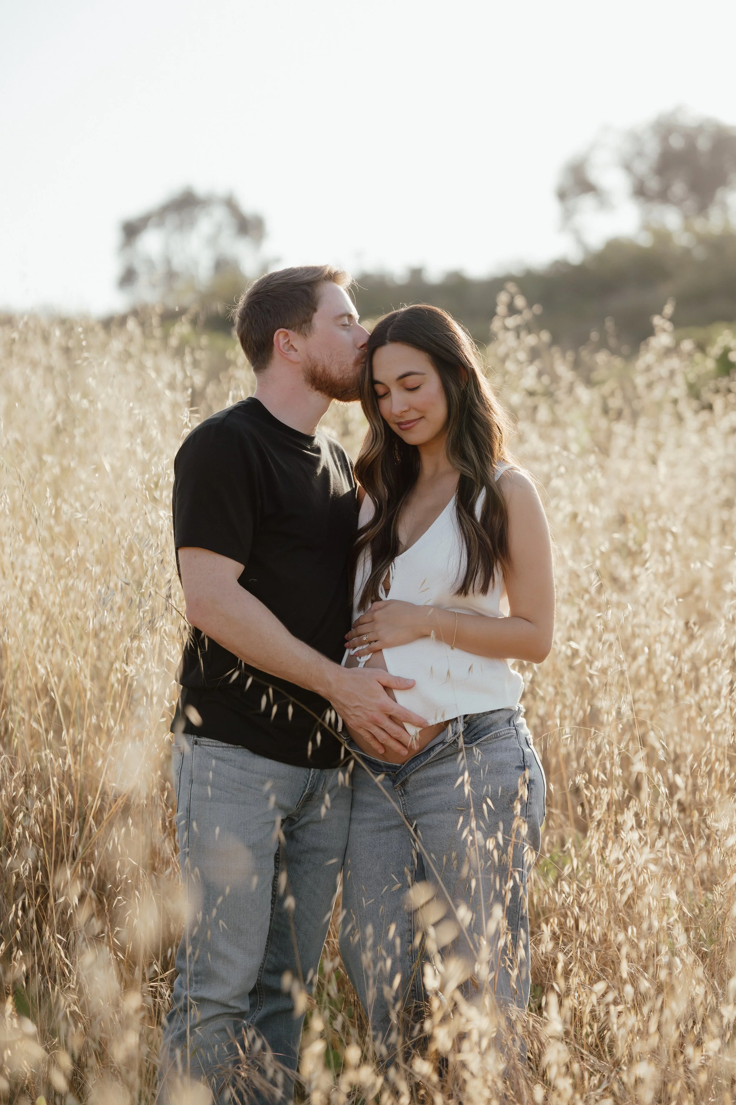 A young man and woman stand close together in a field of tall, golden grass, with the man kissing the woman's forehead and both smiling gently.