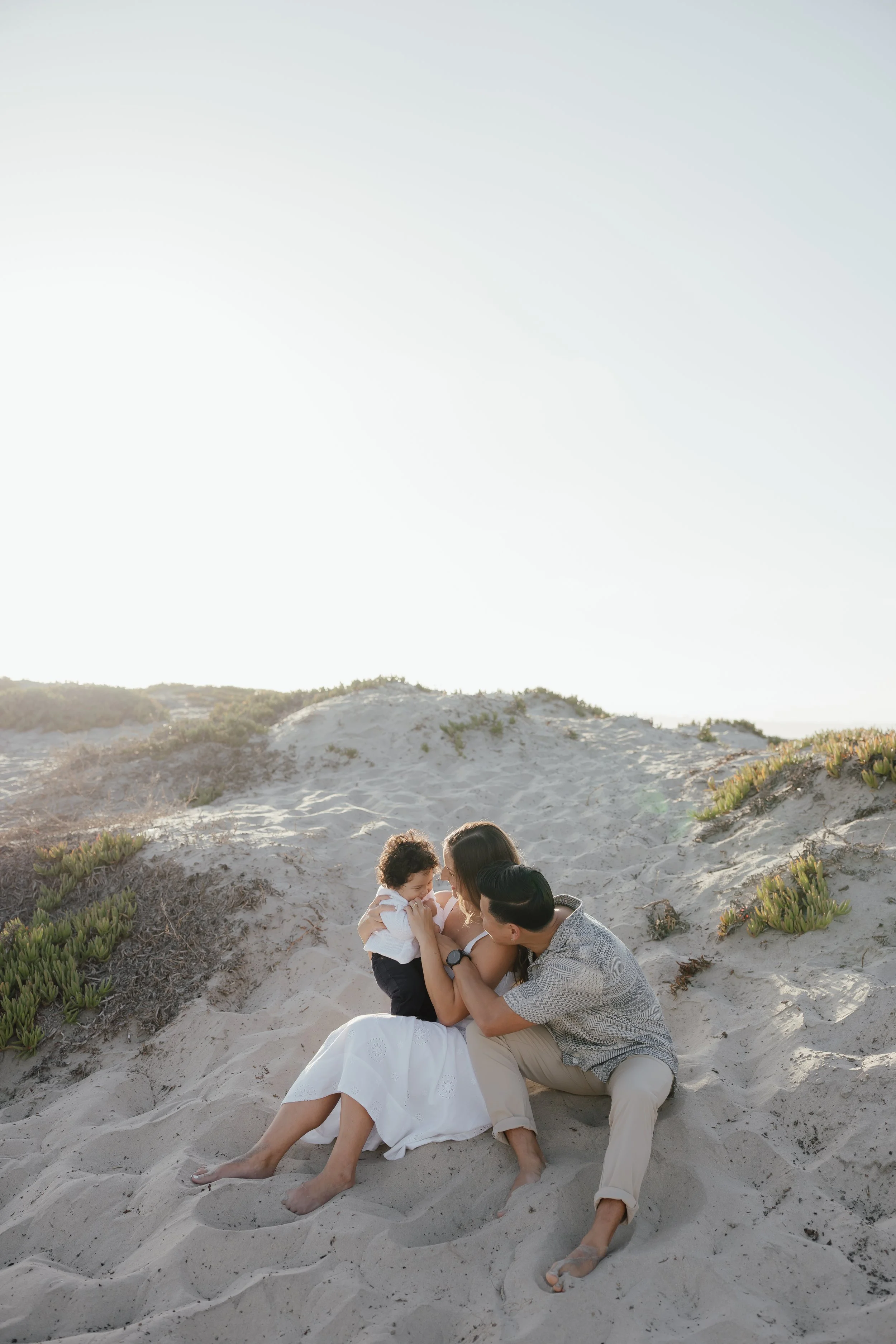 A family of three, a man, woman, and young child, sitting on sandy dunes at the beach, with greenery around, enjoying a sunny day.