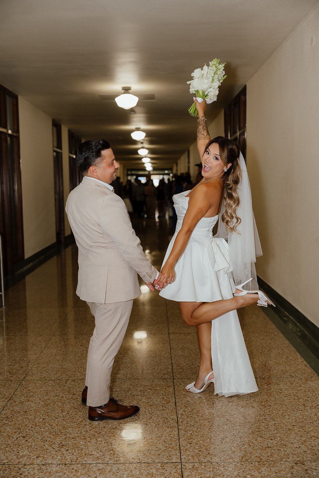 A bride and groom holding hands in a hallway; the bride lifts her foot in excitement while holding a bouquet, and the groom looks at her smiling during San Diego Elopement.