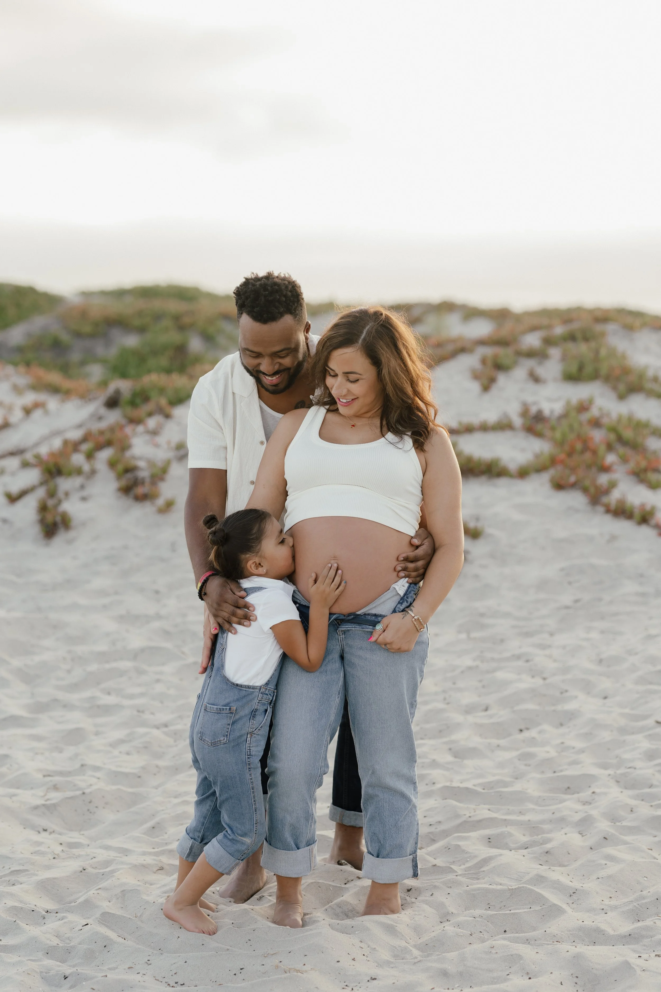 A happy family of three, including a pregnant woman, a man, and a young girl, standing on a sandy beach with dunes and sparse vegetation in the background, smiling and sharing a moment together.