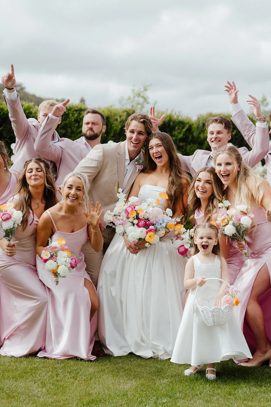 A group of people celebrating a wedding outdoors, including a bride and groom, bridesmaids, groomsmen, and a young girl holding a basket of flowers, all smiling and posing cheerfully at San Diego Wedding.