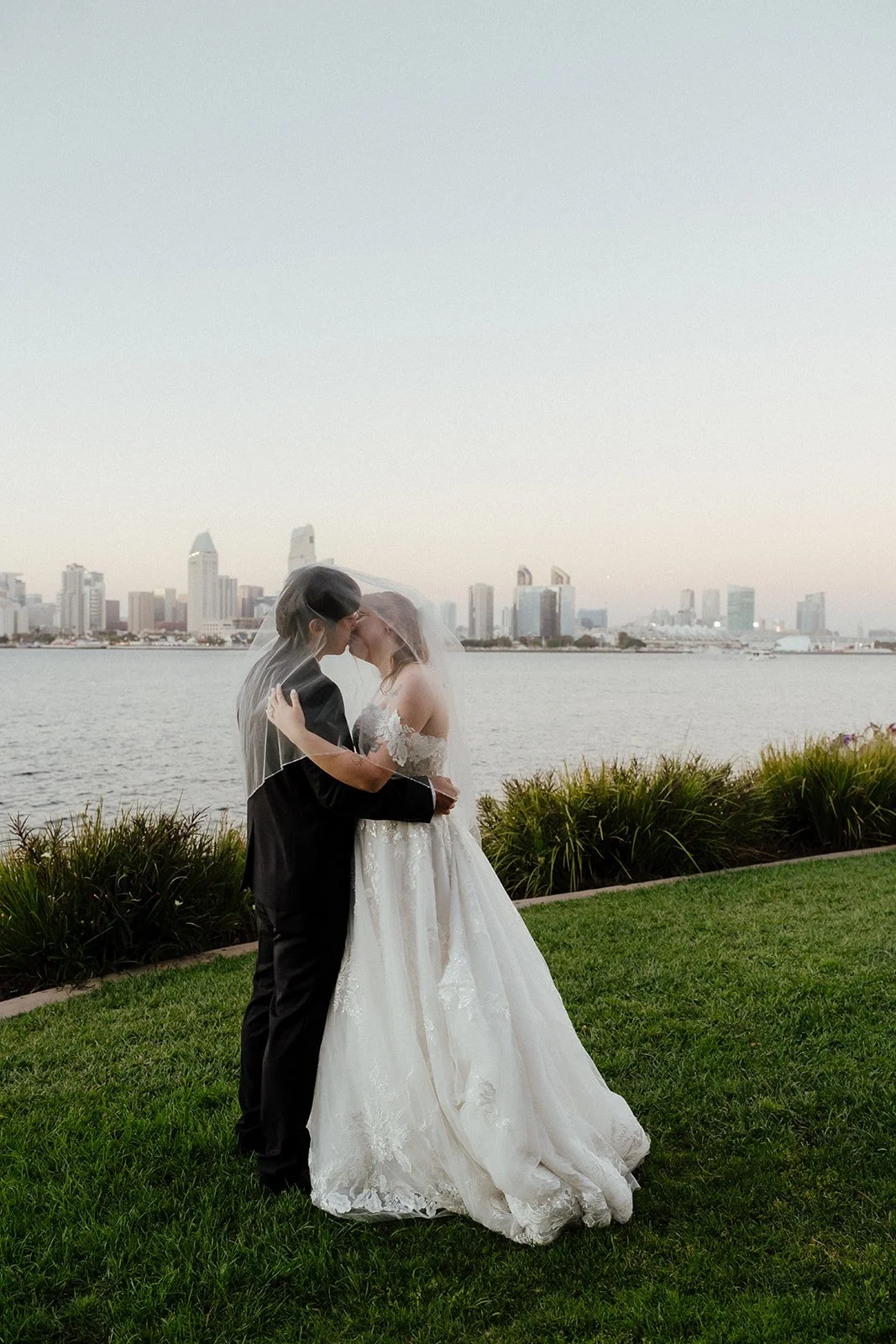 A bride and groom kissing outdoors by water with city skyline in the background, wearing wedding attire, with the bride in a white gown and veil and the groom in a black suit during San Diego Elopement.