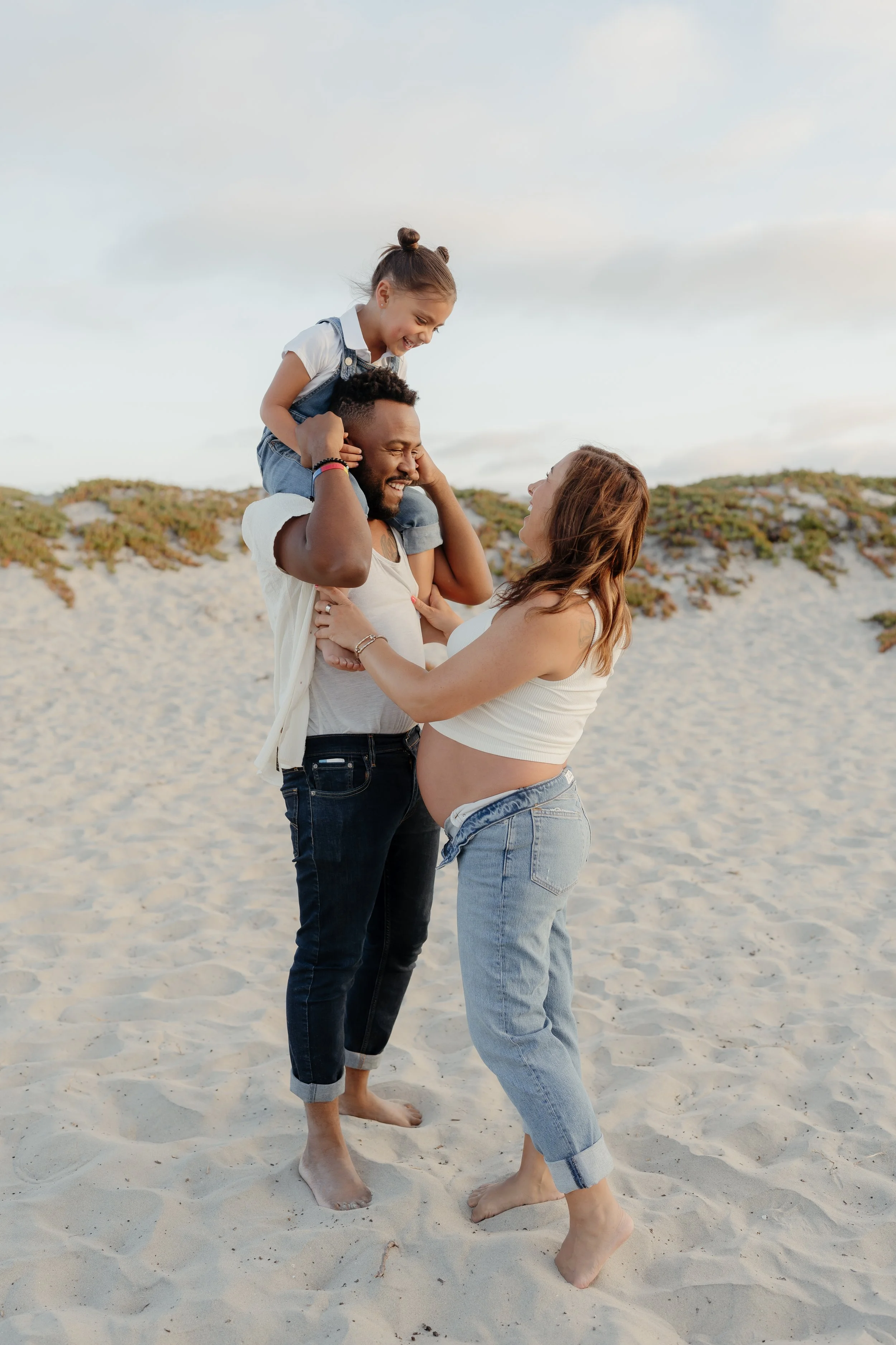 A family of three on a sandy beach, with the mother visibly pregnant, playing and smiling together.