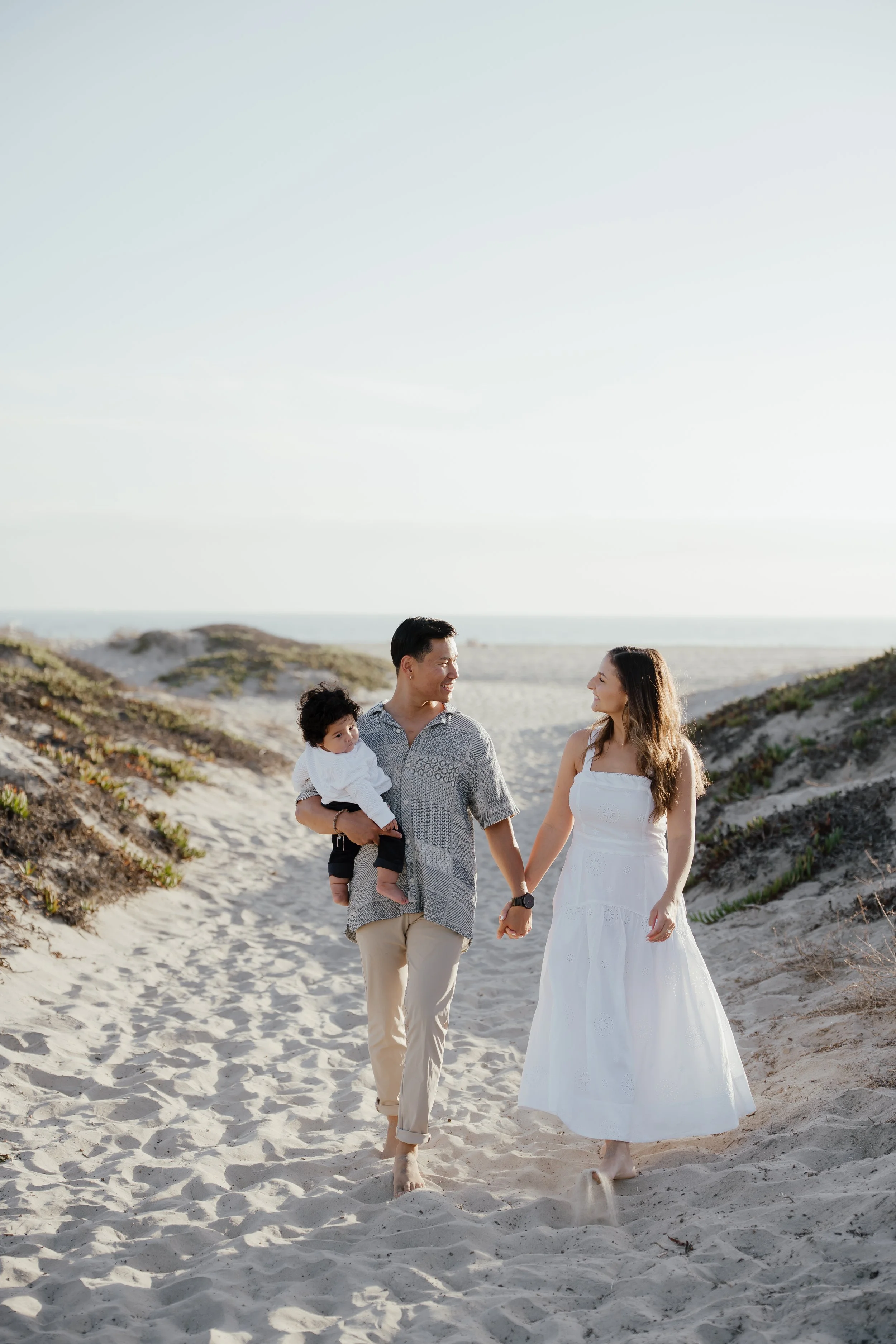 A family of three, including a man, woman, and young child, walking barefoot on the sandy beach holding hands, with dunes on either side and the ocean in the background on a sunny day.