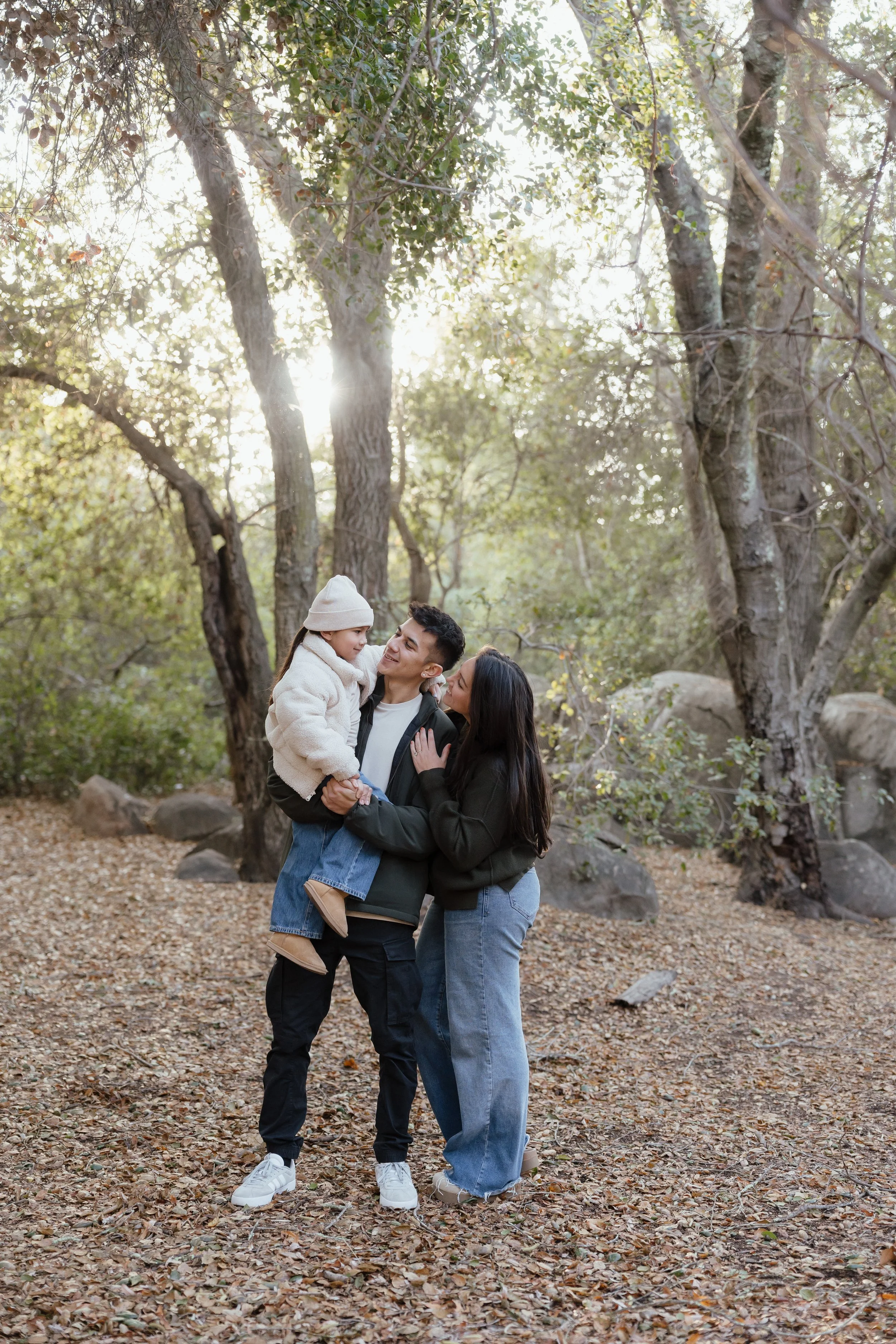 A family of three enjoying time outdoors in a forest during the fall, with sunlight streaming through the trees. The man is holding a young girl, and the woman is standing close beside them, all smiling.
