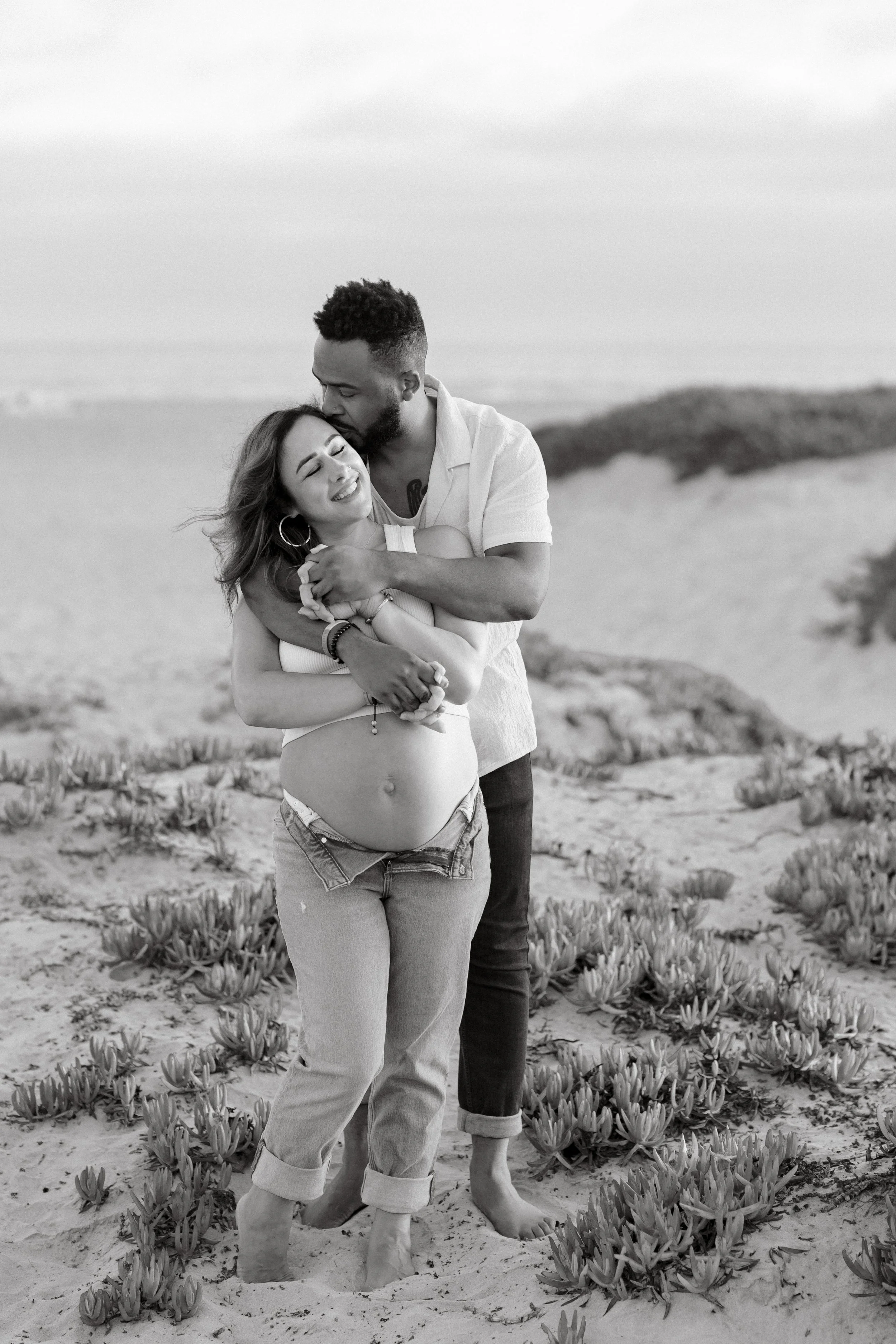 A couple, with the woman being pregnant, embracing and smiling on a sandy beach with plants in the foreground and the ocean in the background.