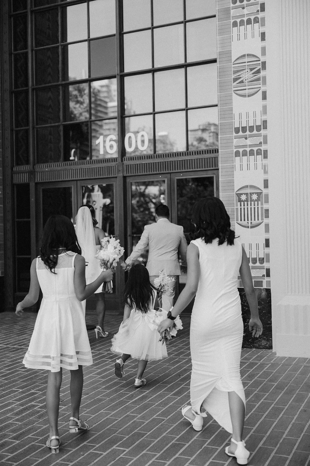 A group of women, a girl, and a man walk into a modern building with glass doors during daytime heading into a Downtown San Diego Elopement.