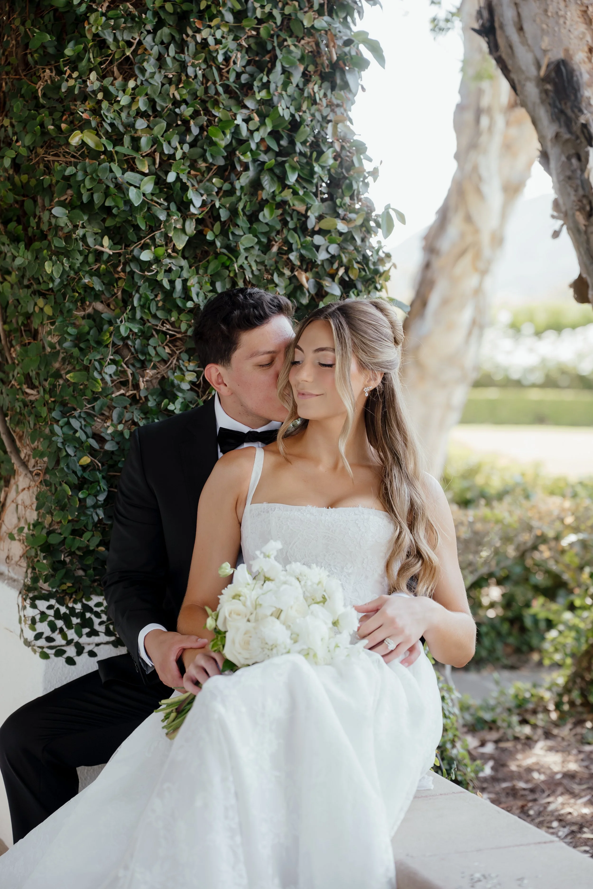 A bride and groom sit closely together outdoors. The bride is holding a bouquet of white flowers. The groom is dressed in a black tuxedo. They are sitting near green bushes and trees, sharing a tender moment at Twin Oaks Gold Course San Diego.