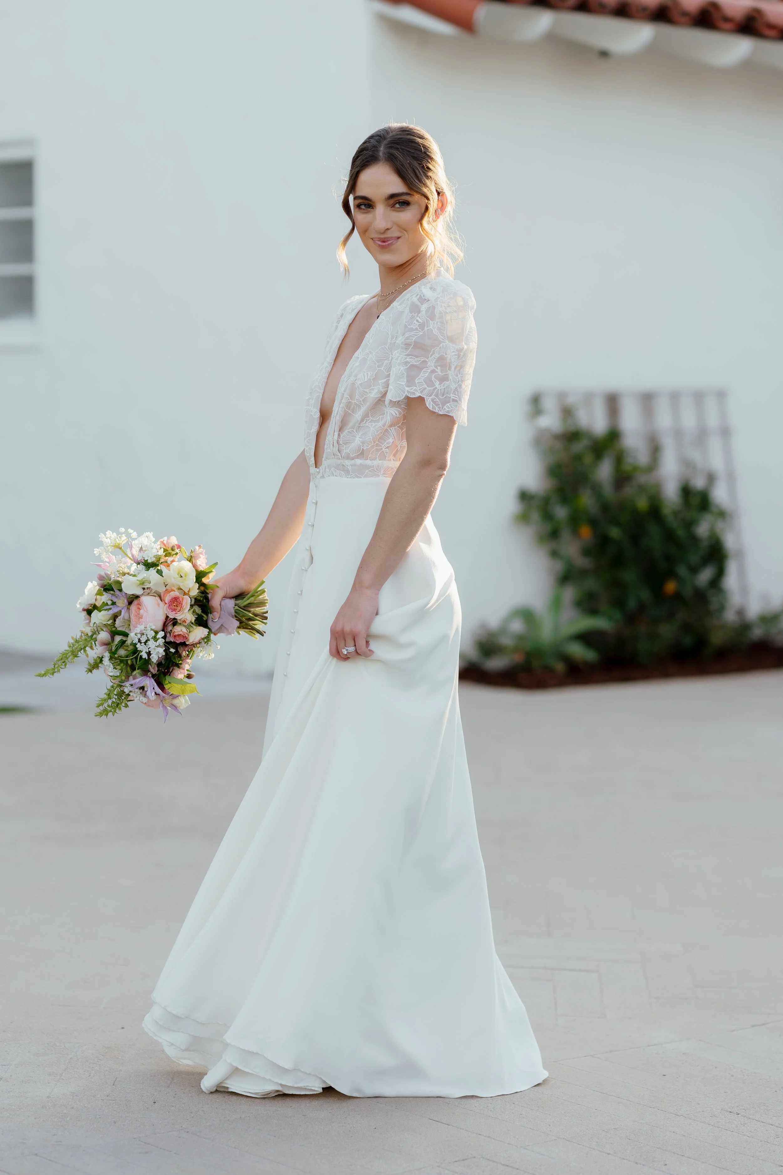 A woman in a white wedding dress holding a bouquet of pink and white flowers, standing outdoors in front of a white wall with greenery.