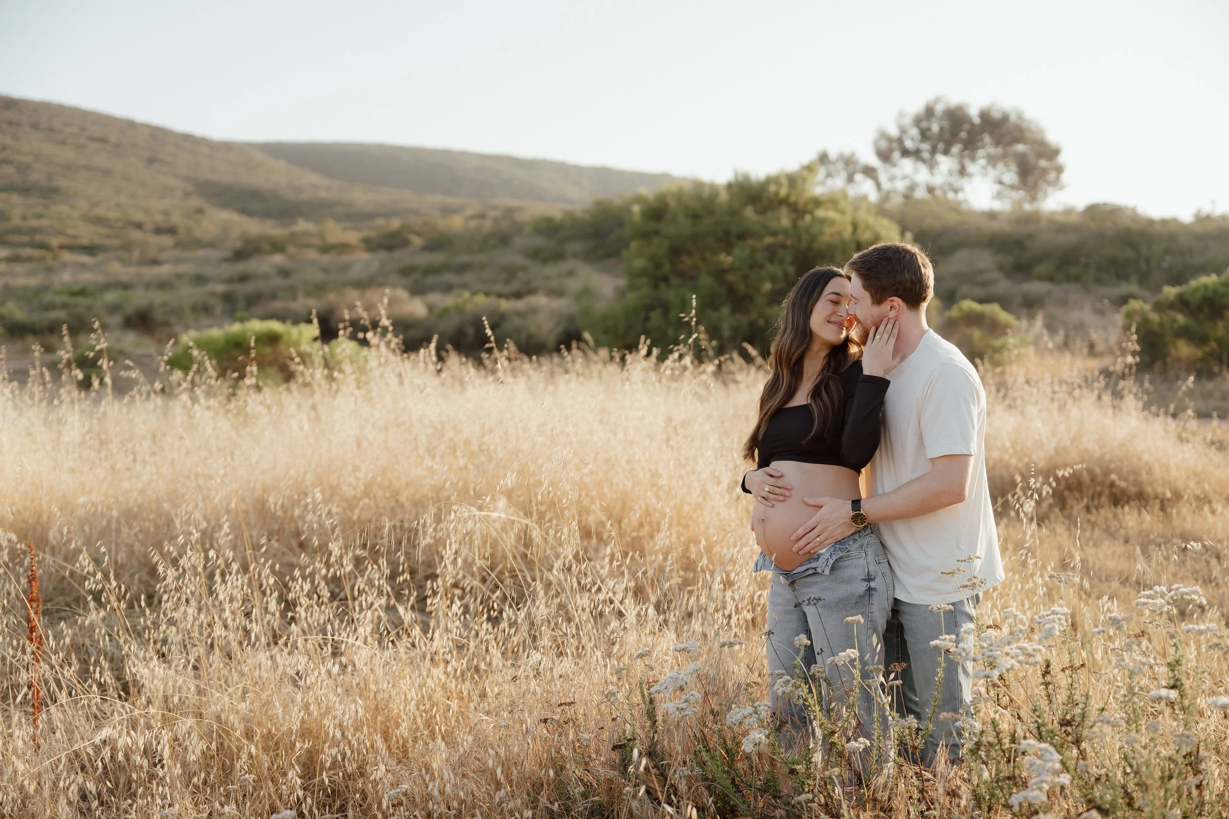 A couple in a field of tall, dry grass, embracing and smiling at each other, with hills and trees in the background during late afternoon or early evening.