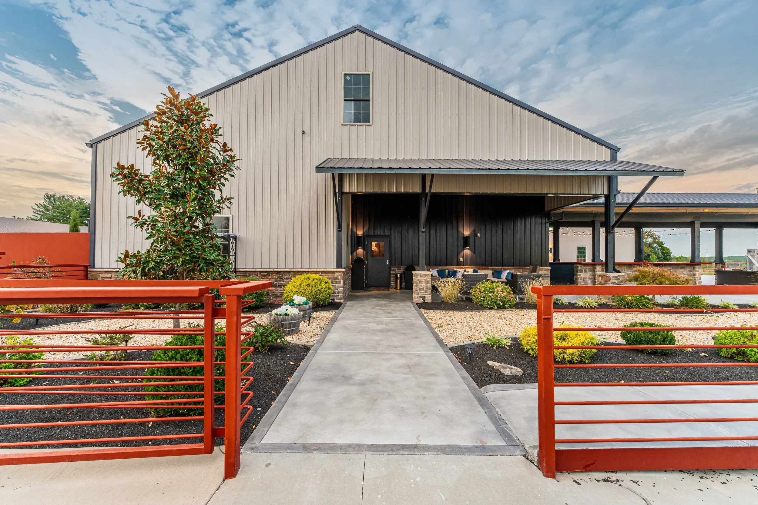 A modern barn-style building with vertical siding, a small covered porch area, and a pathway leading to the entrance. The front yard features a landscaped garden with bushes, flowers, and a small tree, enclosed by a red metal fence. The sky is partly cloudy with a peaceful ambiance.