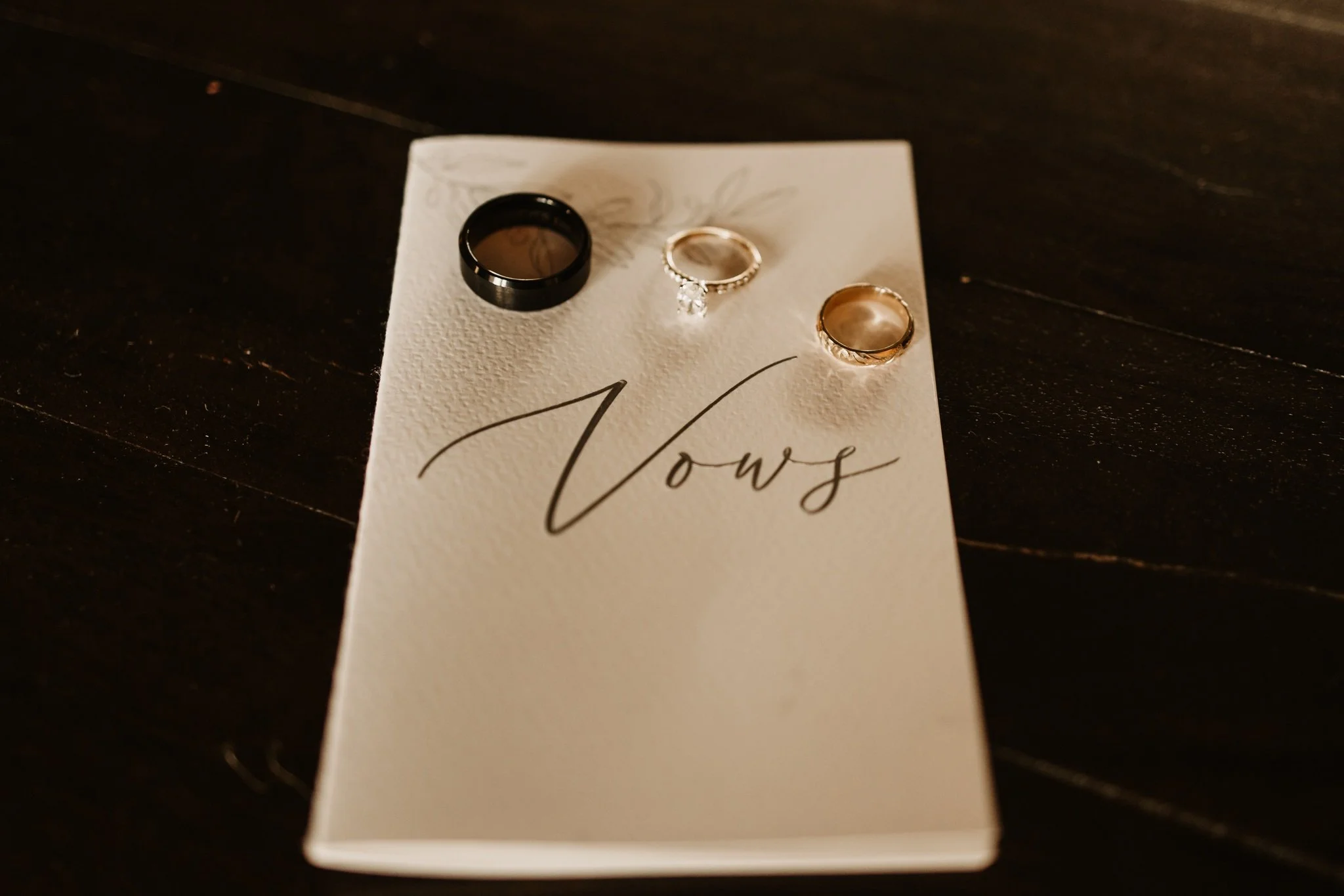 Black and gold wedding bands along with an engagement ring with a clear diamond, placed on a handwritten note that reads 'Thanks', on a dark wooden surface.