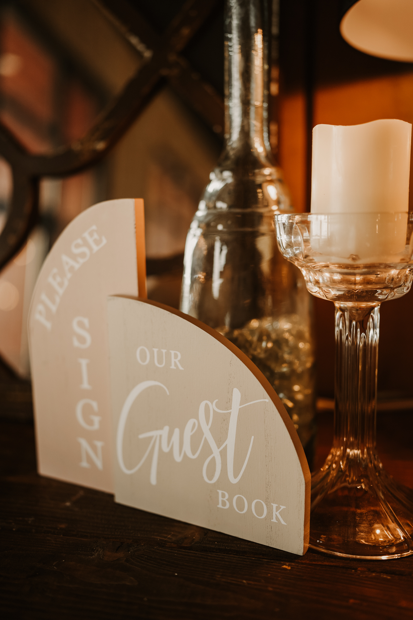 Close-up of a decorative candle on a glass holder, a silver vase, and a sign with text on a dark wooden surface, in a cozy, warmly-lit setting.