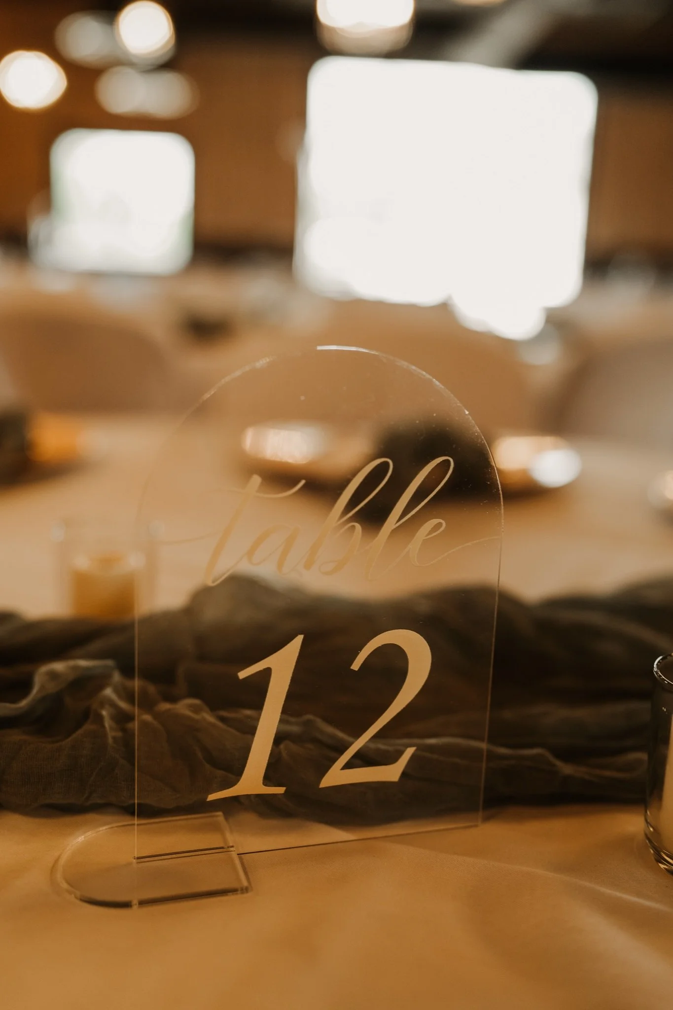 Close-up of a glass decoration with the word 'Celebrate' and the number '12' written on it, placed on a table with a dark brown cloth and a blurred background of warm indoor lighting.