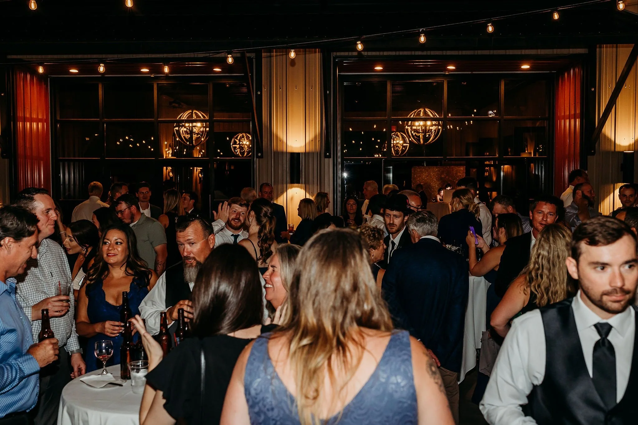 A crowded indoor party or social event with many people socializing, holding drinks, and enjoying the evening. The room features warm lighting, visible through large windows, and hanging light bulbs adding to the ambiance.