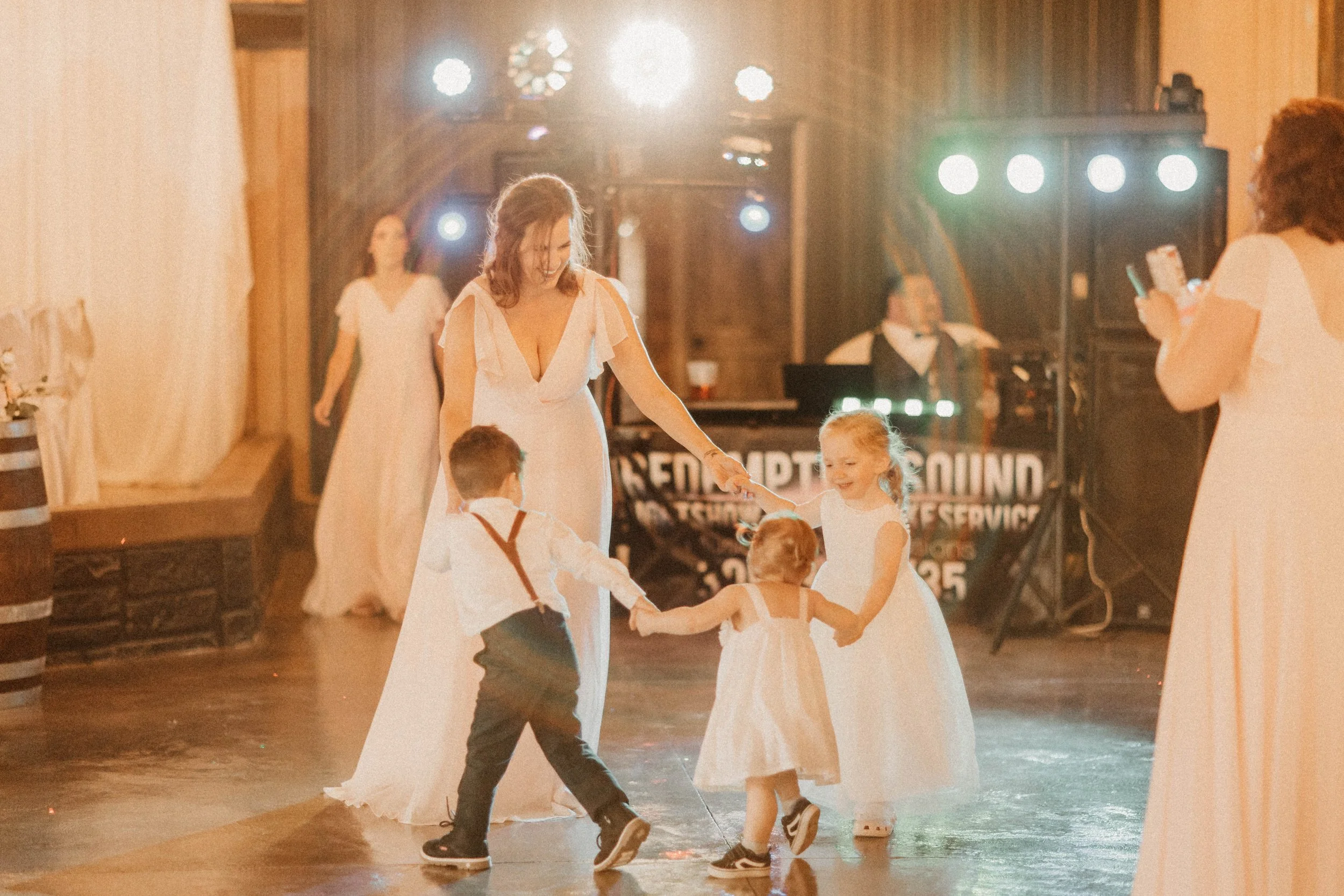 Children and a woman dance together in a circle at an indoor event with warm lighting and a DJ in the background.
