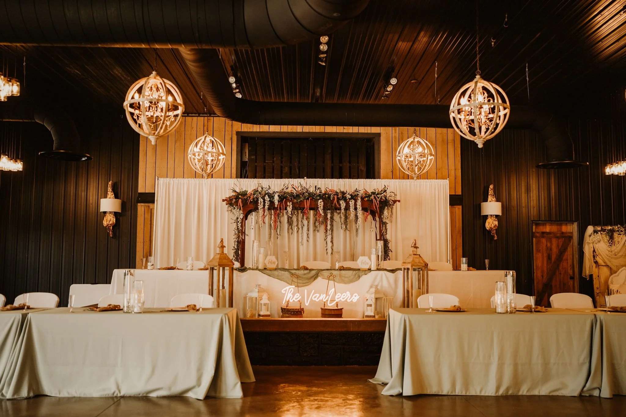 Wedding reception setup with long tables covered in white tablecloths, decorated with small lanterns and dried flowers. The background features a floral wedding arch and a backdrop with the words 'The Vanels' in cursive lights, under warm hanging lig