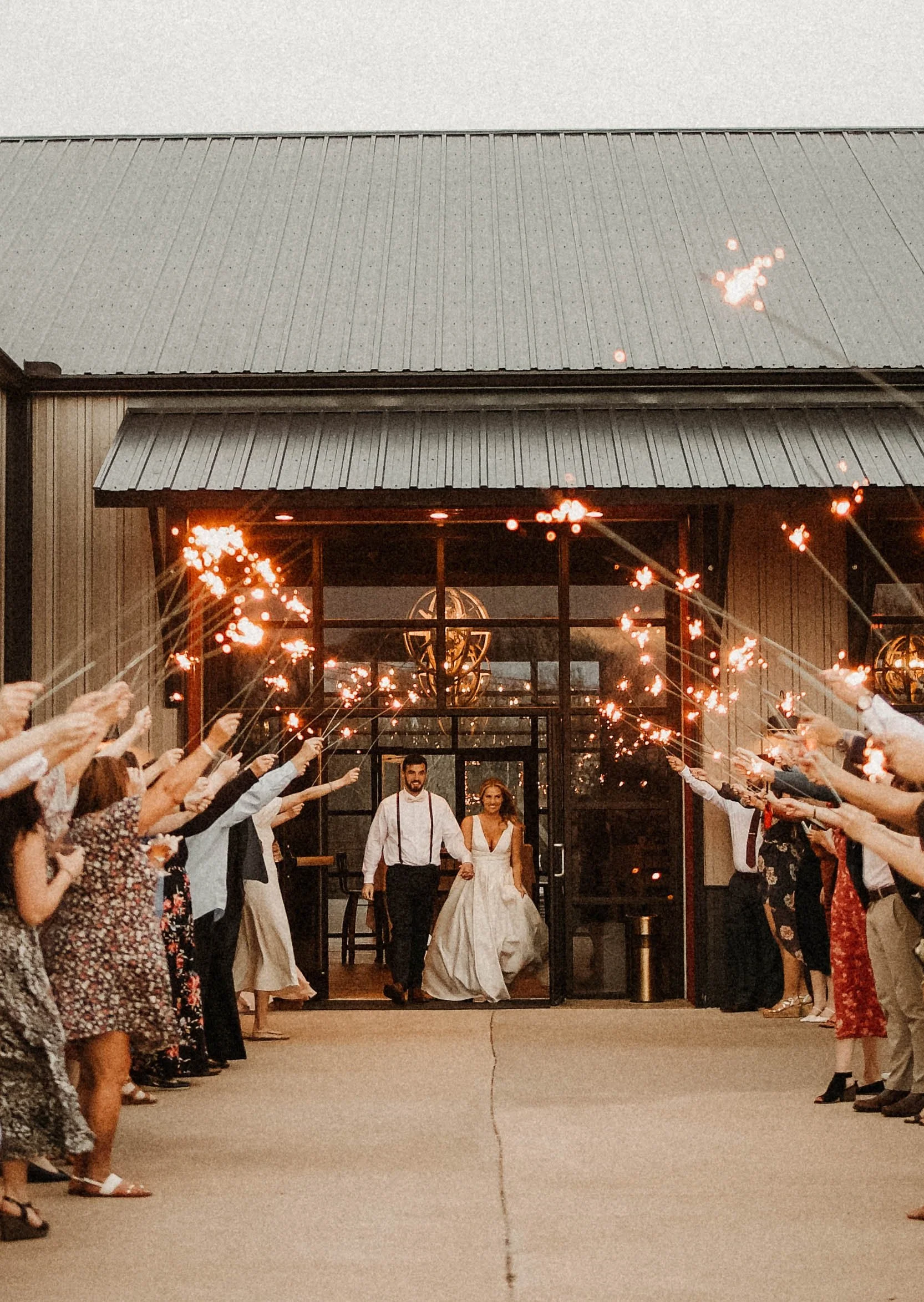 A bride and groom walking out of a building, holding hands, as guests line up on either side holding sparklers, celebrating their wedding.