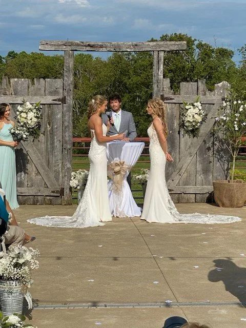 Couple getting married outdoors under a wooden arch, with bridesmaids and floral decorations.