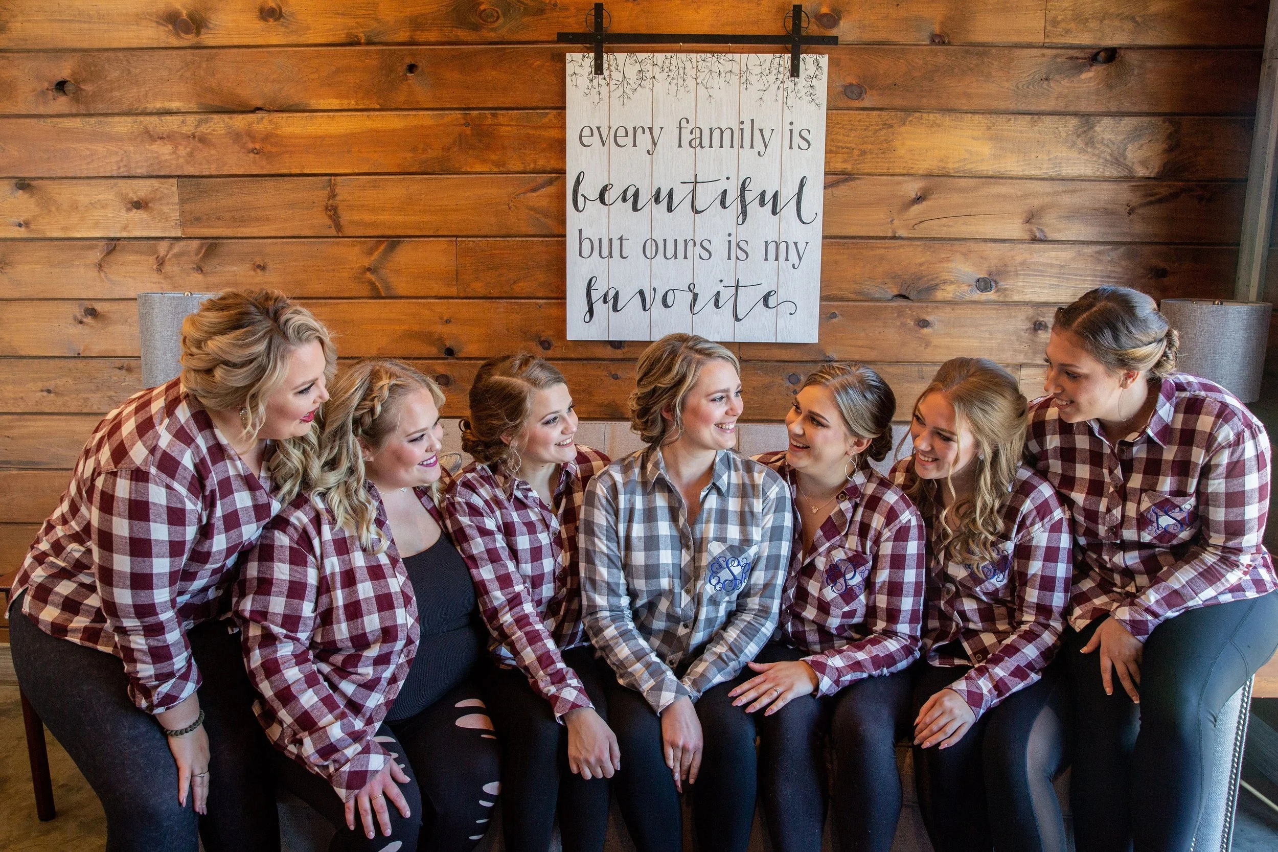 A group of seven women sitting closely together, wearing matching red and black plaid shirts, smiling and talking in a rustic wooden room with a wall hanging that reads 'every family is beautiful but ours is my favorite'.