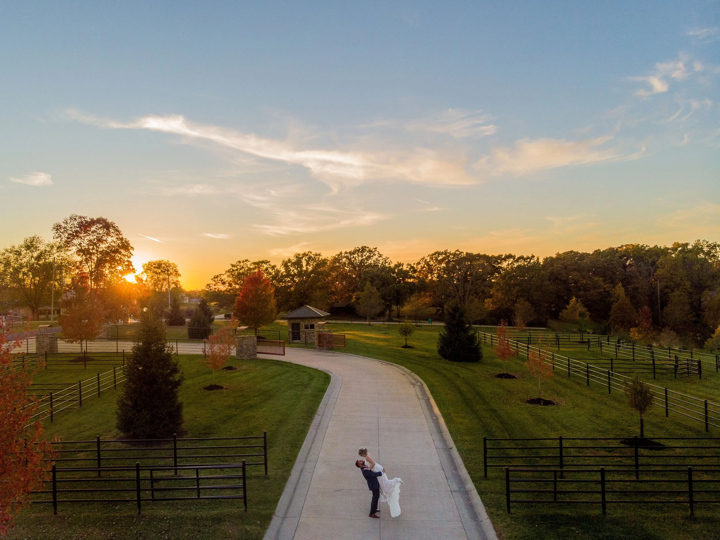 A newlywed couple shares a kiss in a park during sunset with a paved walkway, trees, and a small building in the background.