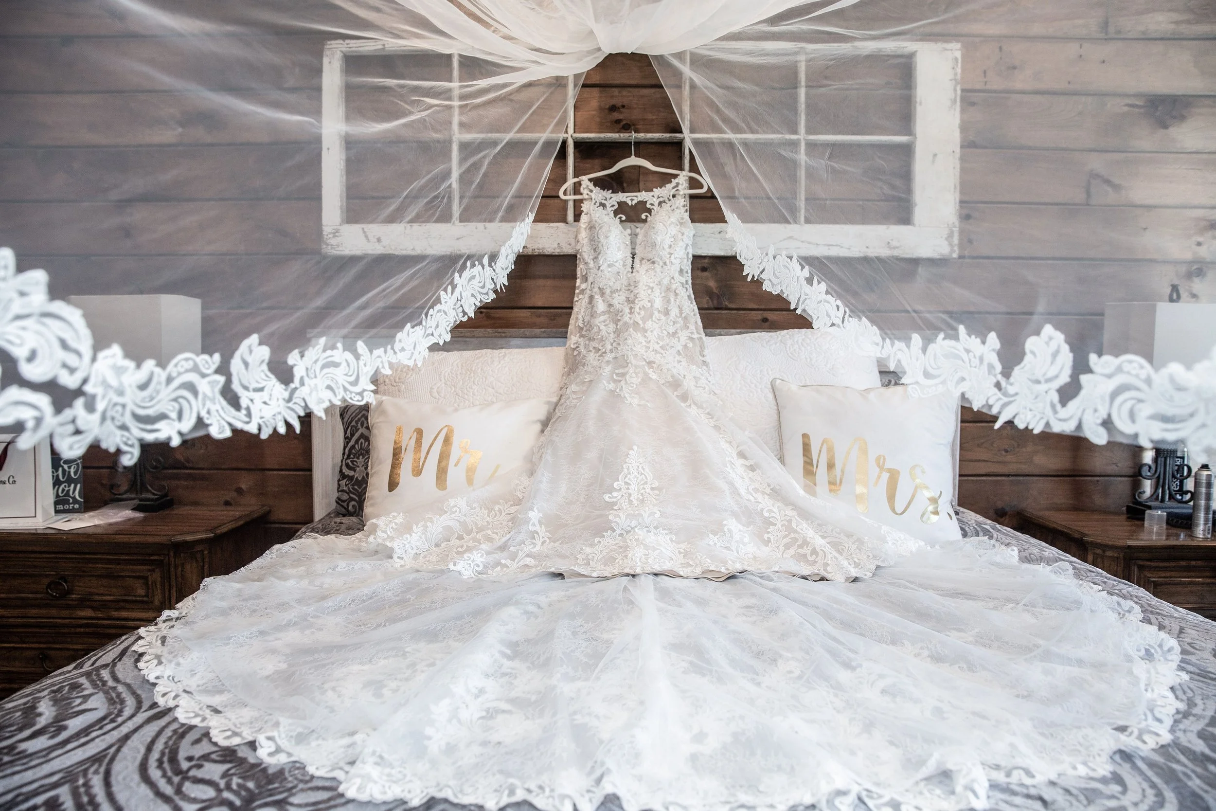 A wedding dress hanging on a white hanger under a sheer canopy over a bed with pillows that say 'Mr.' and 'Mrs.'