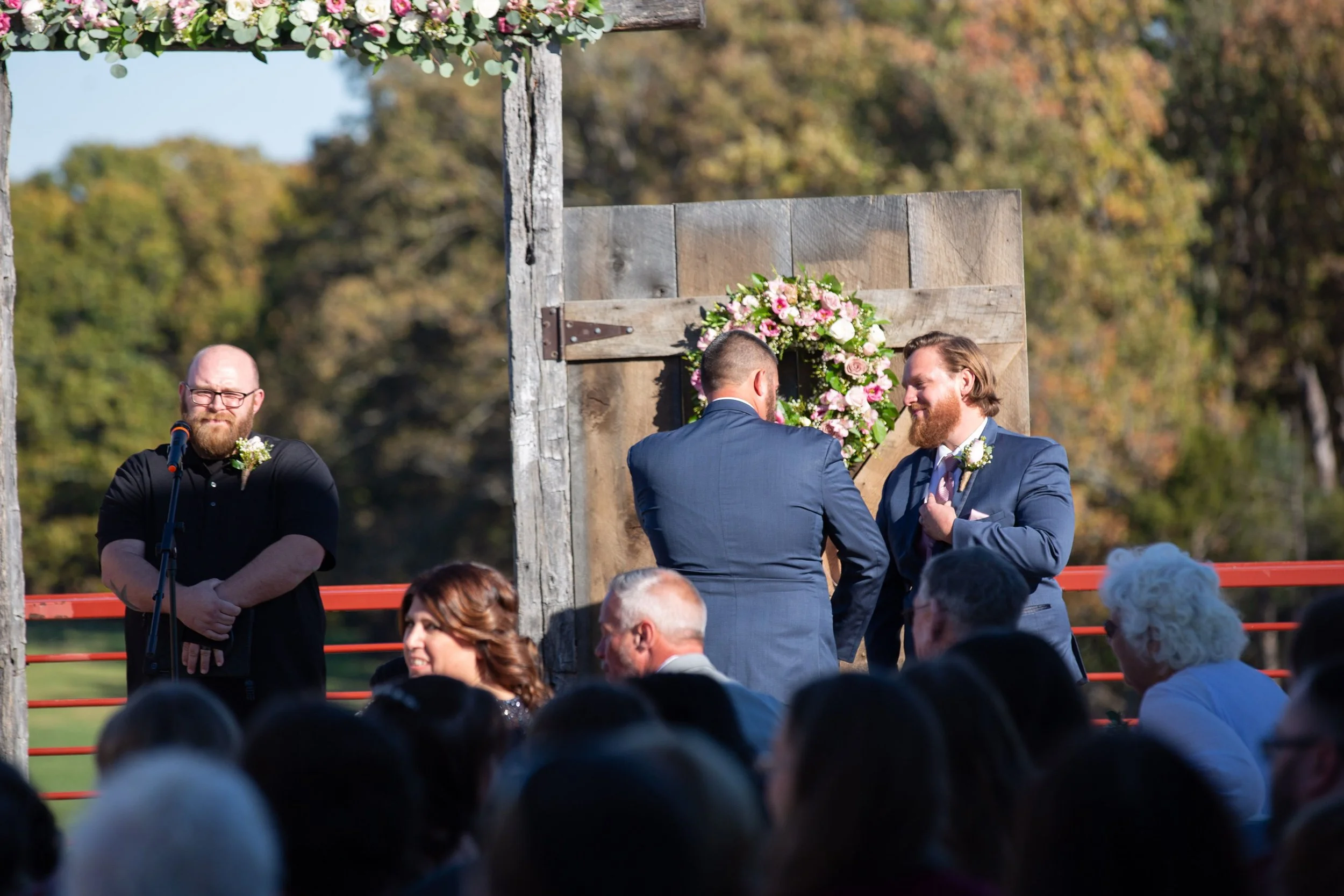 A wedding ceremony outdoors with two grooms exchanging rings in front of a floral wreath. The scene includes a officiant and guests, with trees and a wooden backdrop.
