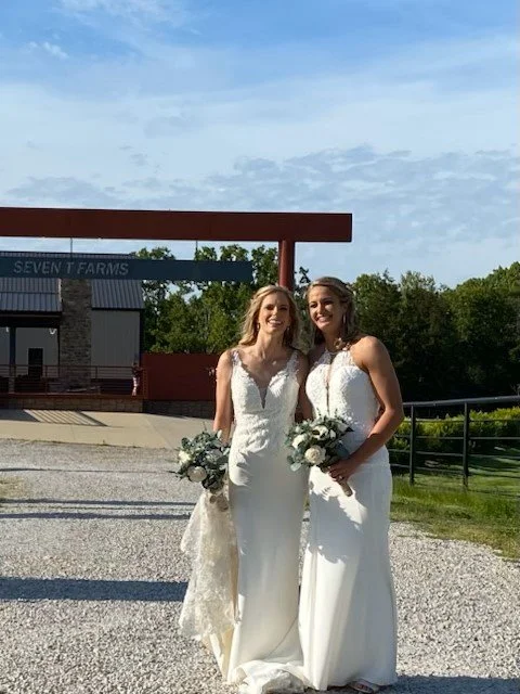 Two women in wedding dresses holding bouquets, smiling outdoors in front of a building with a sign reading "Seven T Farms".