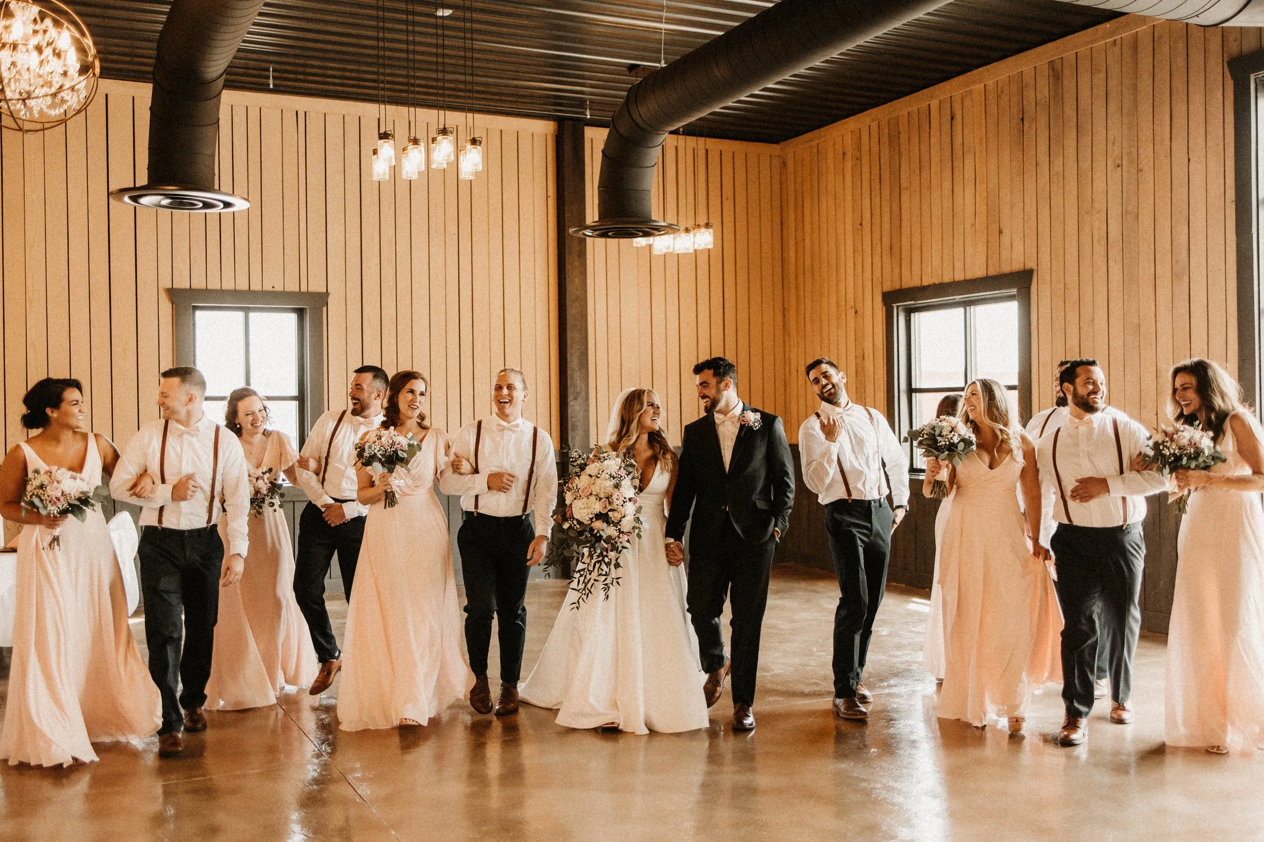 A wedding party dancing indoors with wooden walls, large windows, and warm lighting.