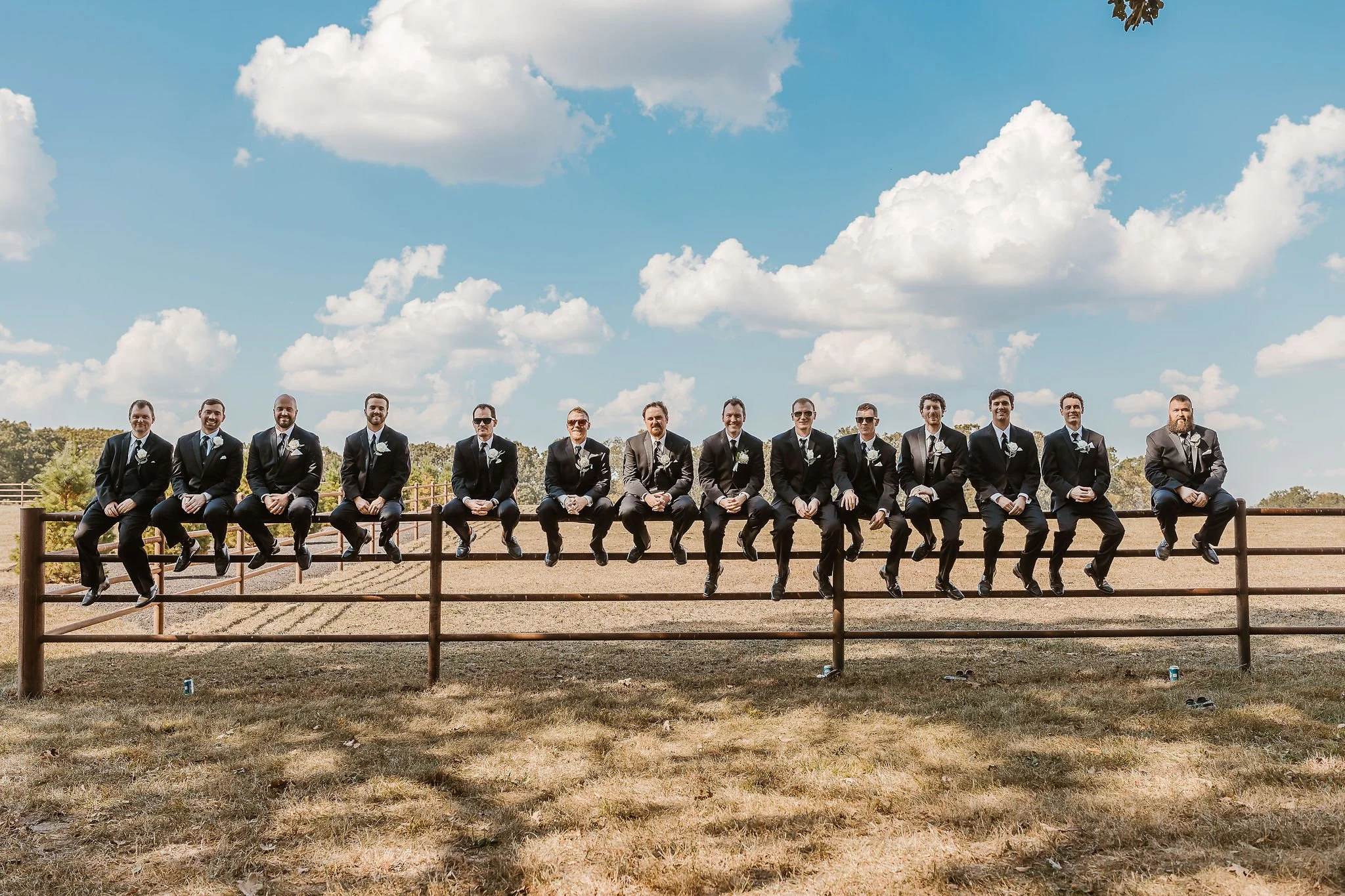 A group of men dressed in black tuxedos with white shirts and black bow ties, sitting on a wooden fence outdoors under a blue sky with scattered white clouds.