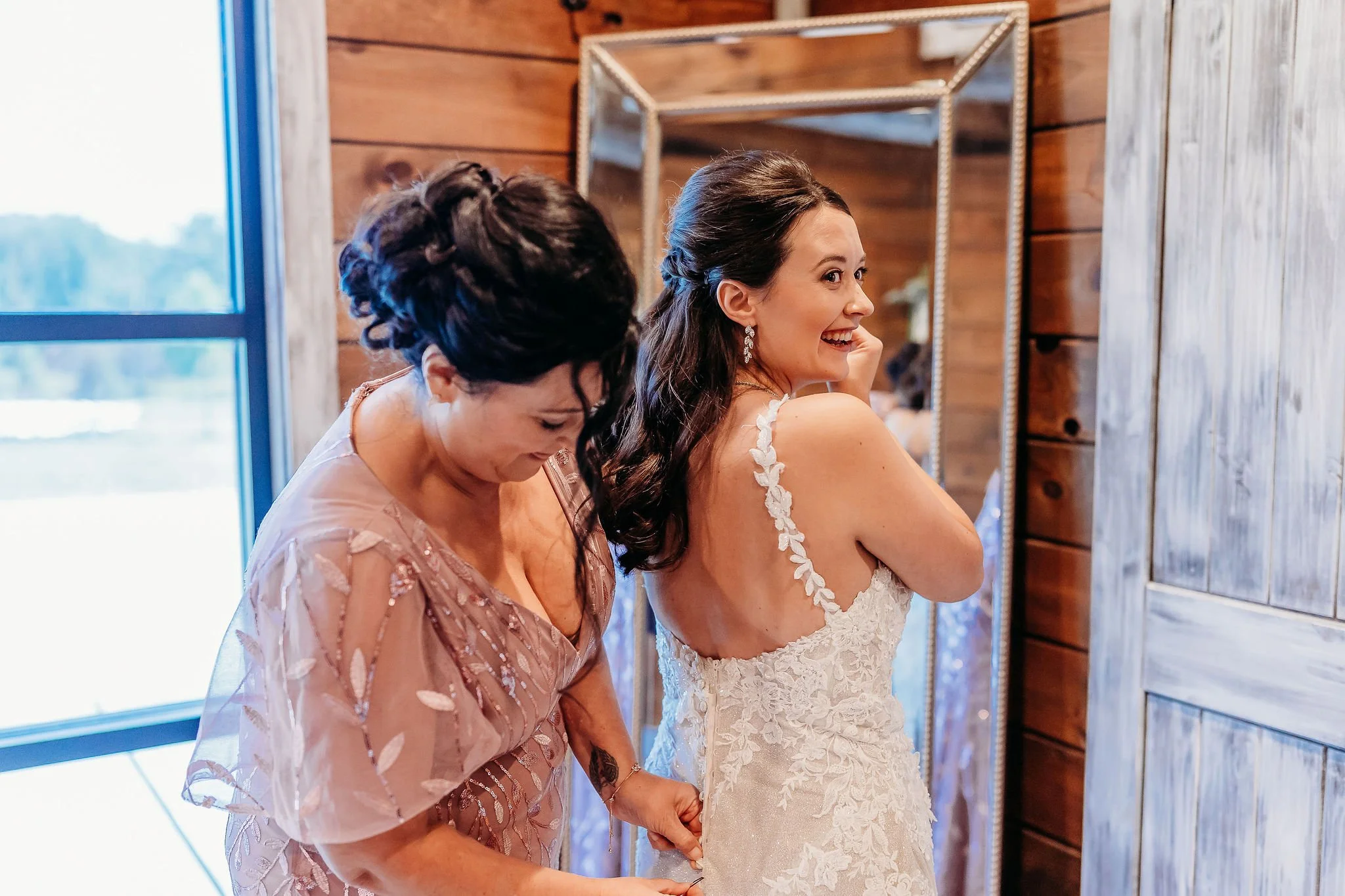 A bride smiling and laughing while in her wedding dress, with a woman in a pink dress helping her, indoors next to a mirror and wooden wall.