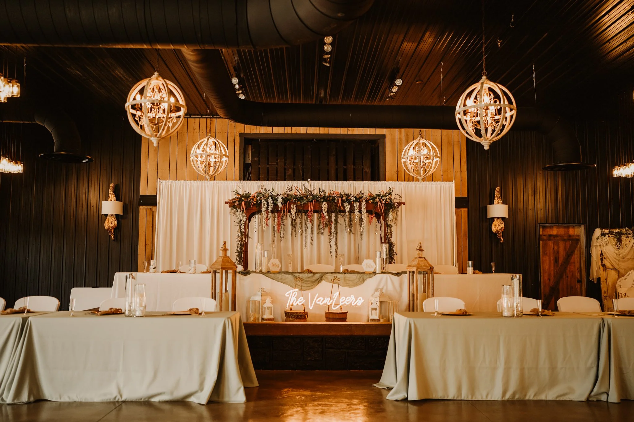 Wedding reception area with a decorated head table, floral arrangements, lanterns, and a backdrop with the words 'The Van Leeers,' inside a rustic-style venue with warm lighting and wood-paneled ceiling.