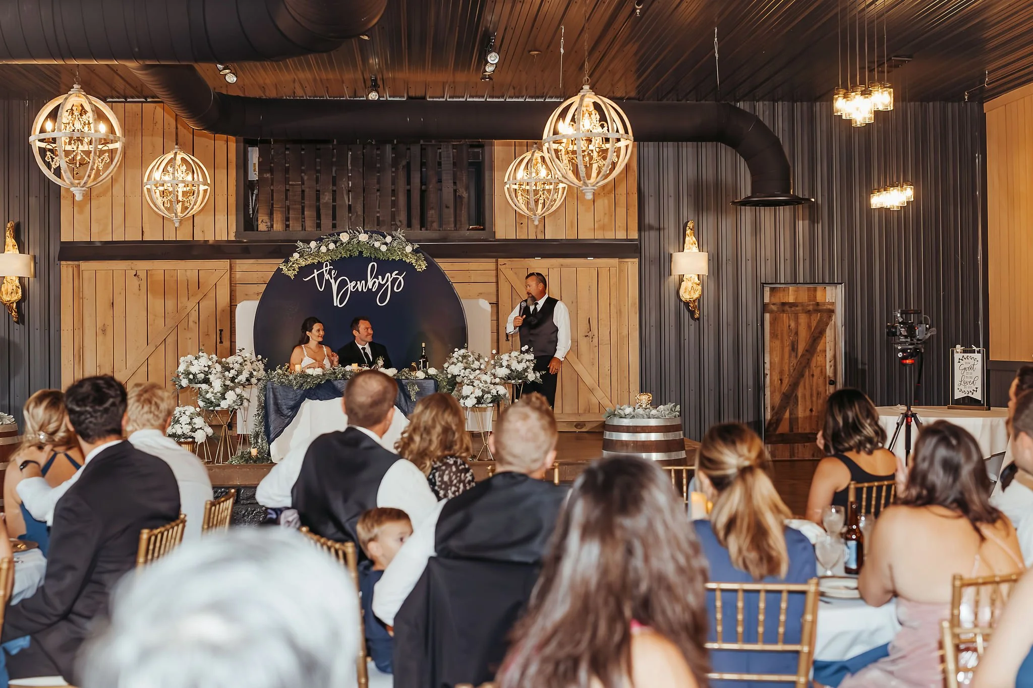 A wedding reception in a rustic barn with wooden walls and black paneling. The bride and groom are seated at the front table decorated with white flowers, with a black backdrop that says 'the Denbys'. A man in a vest and tie is speaking into a microp