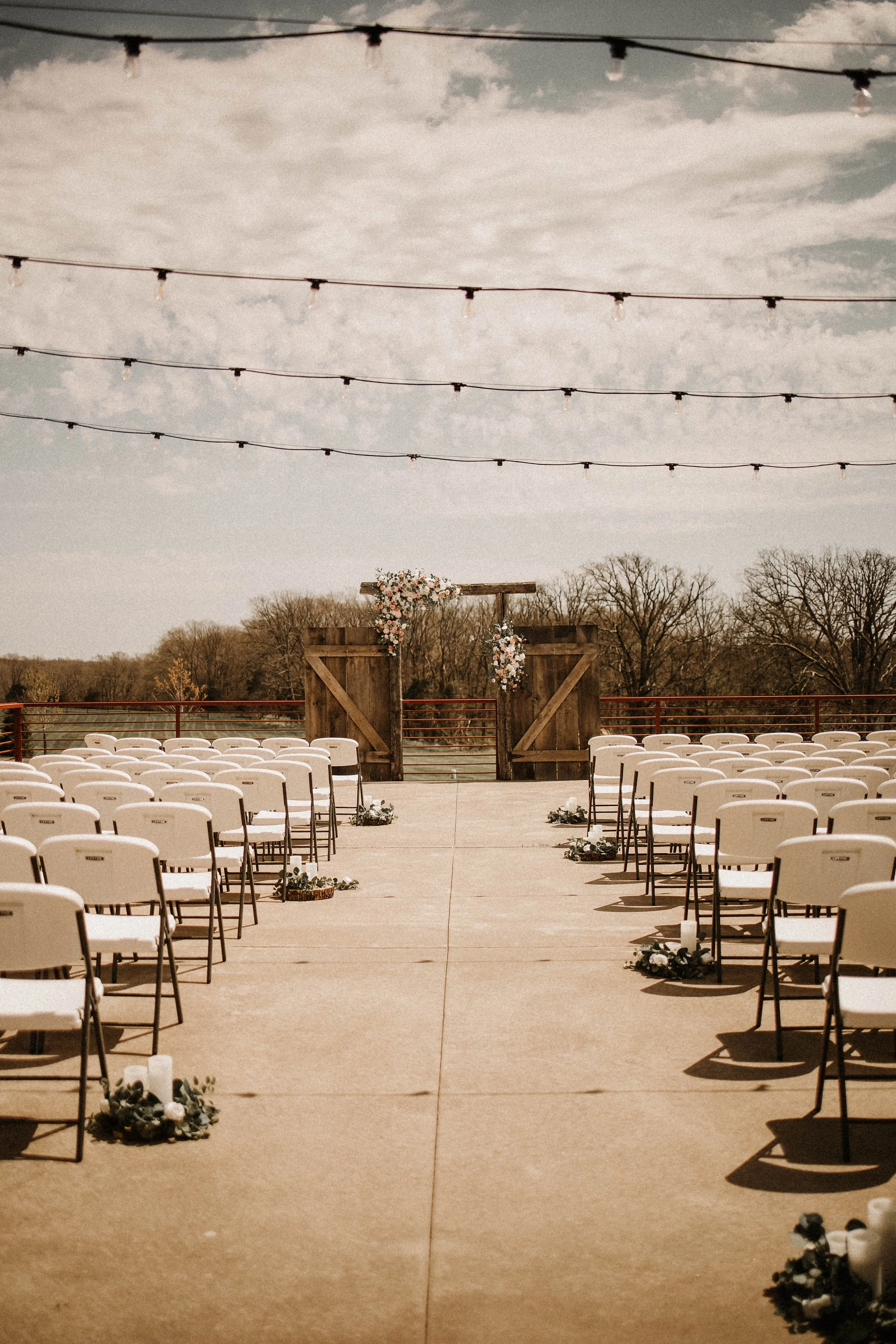 Outdoor wedding ceremony setup with rows of white chairs, floral arrangements, a wooden arch decorated with flowers, and string lights hanging overhead, with a lake and trees in the background.