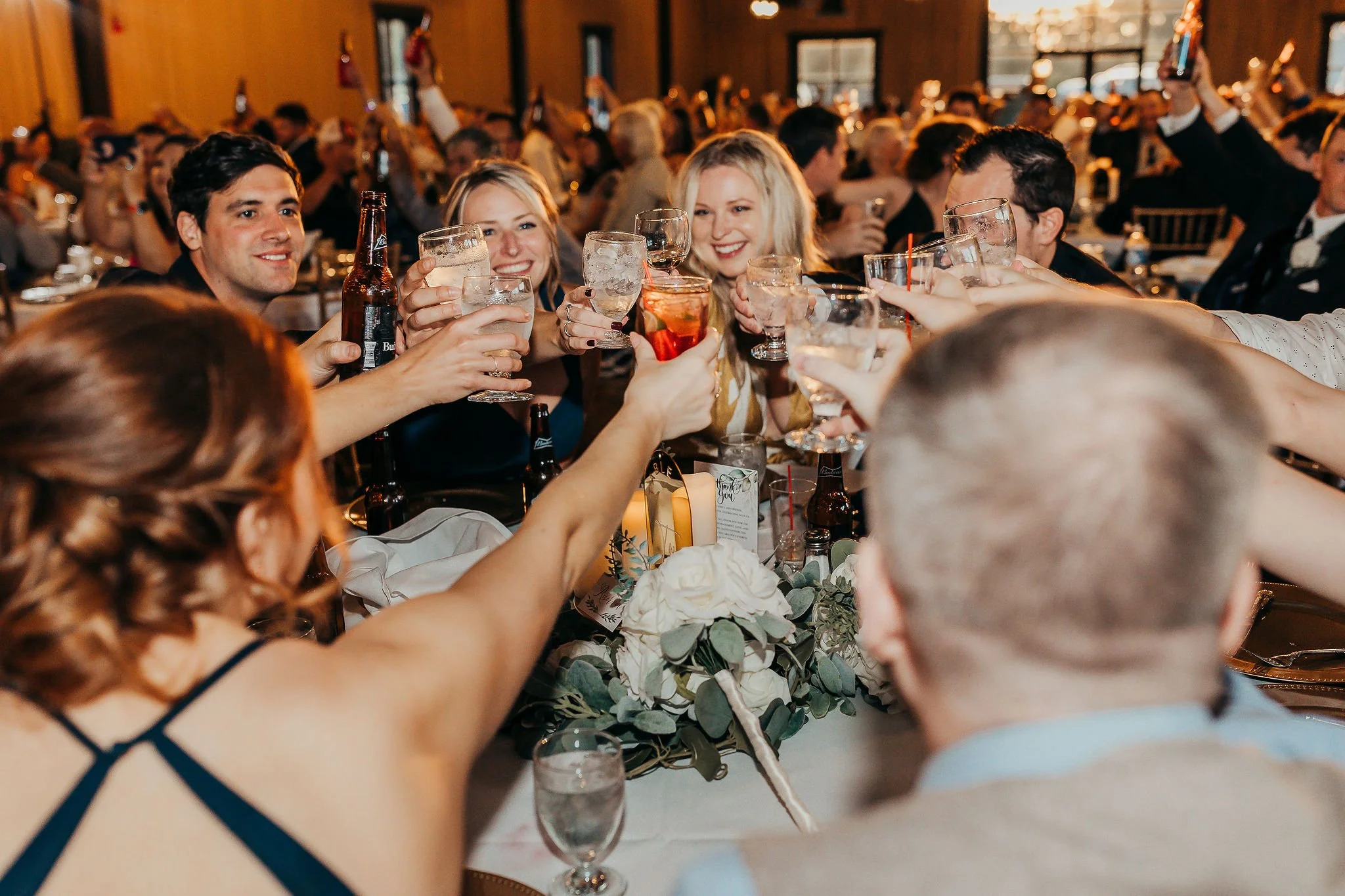 People raising glasses in a toast at a celebration or party in a crowded indoor venue with warm lighting and floral centerpiece on the table.