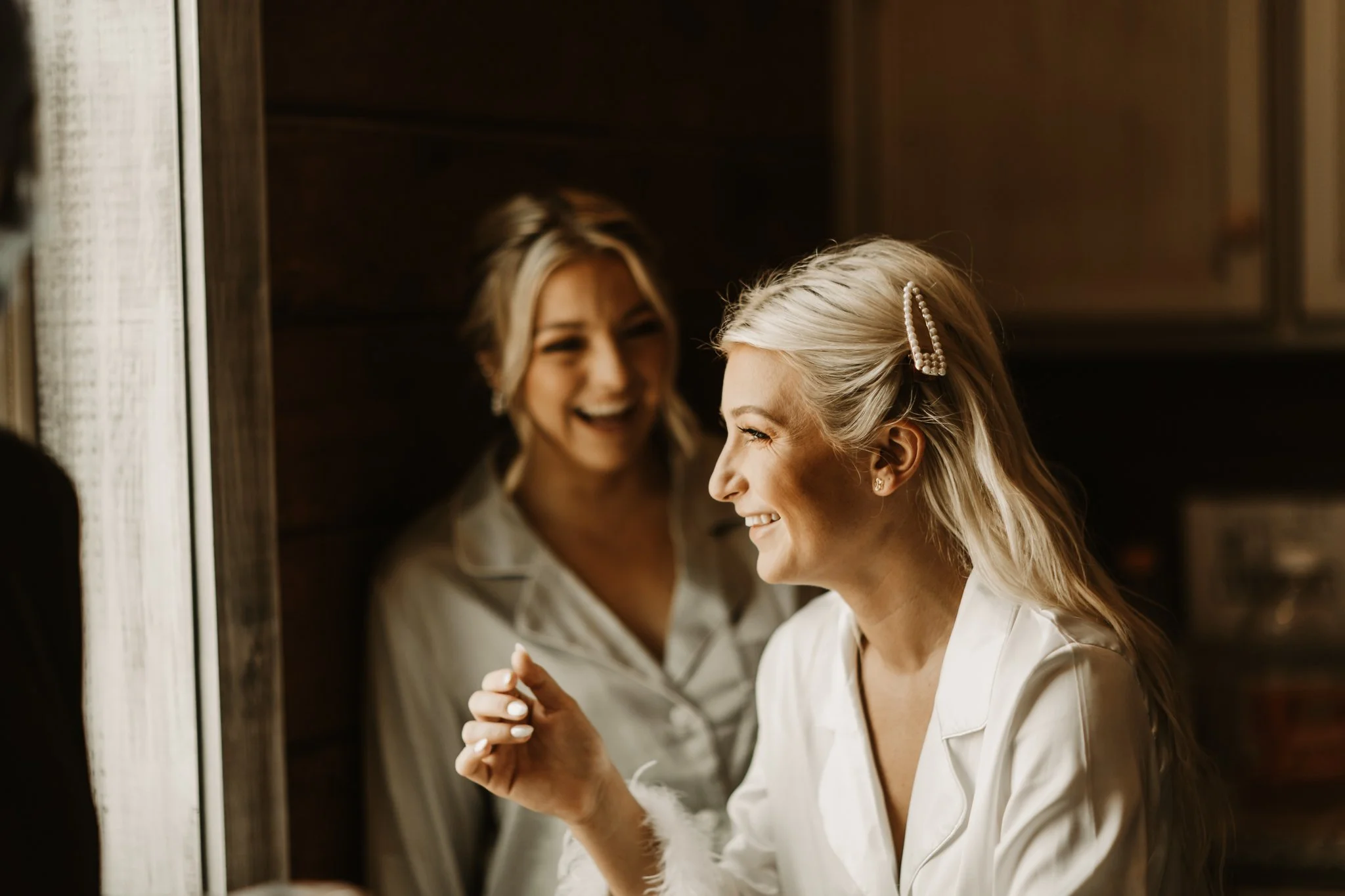 A woman with blonde hair styled with a pearl hair clip is smiling as she looks at herself in a mirror, with another woman also smiling in the background.