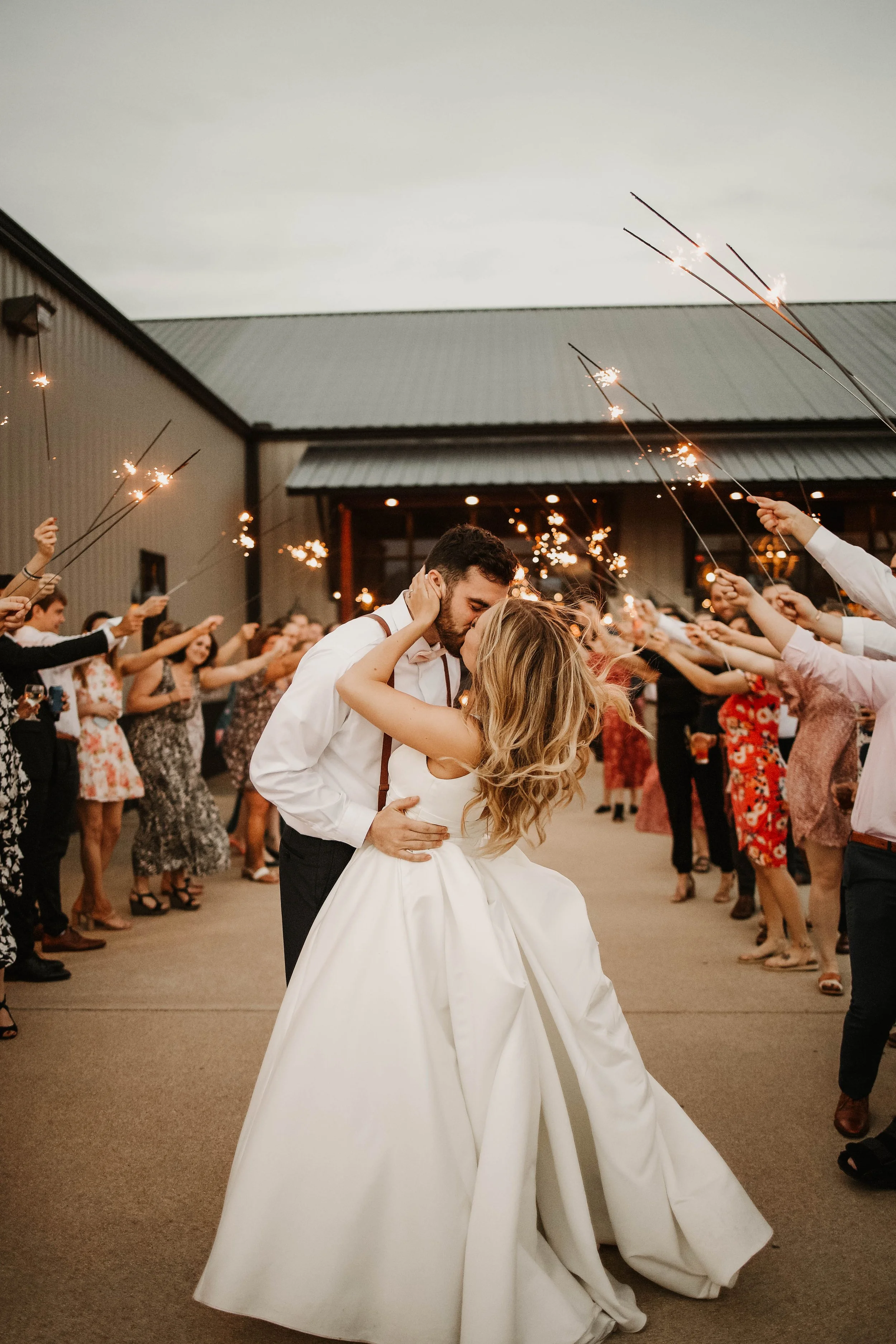 A bride and groom share a kiss while dancing at their wedding reception, surrounded by guests holding sparklers.