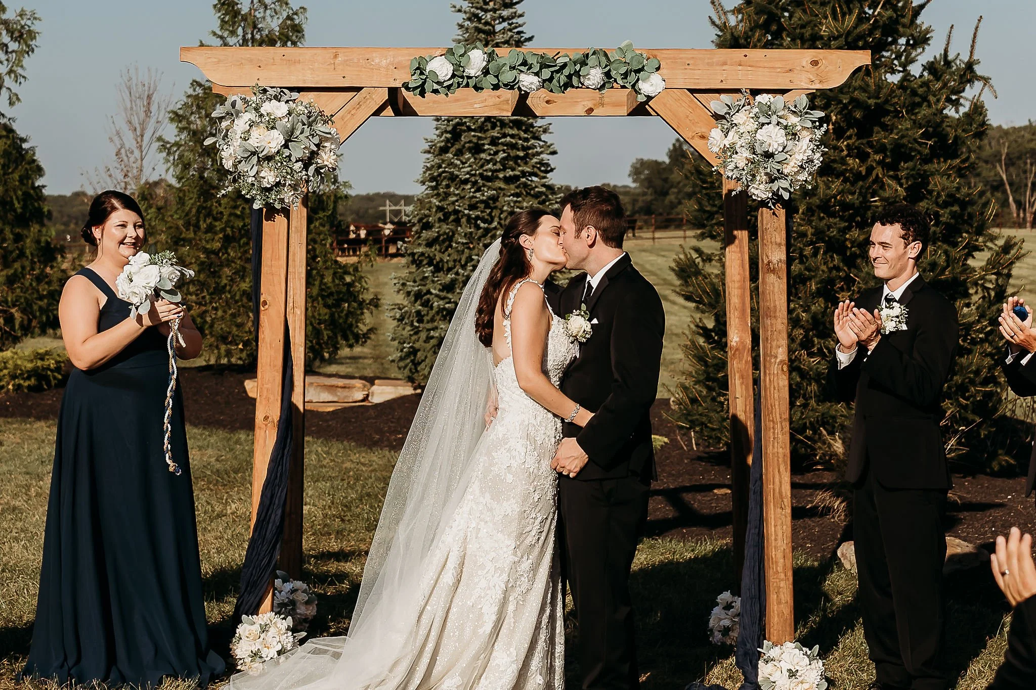 A bride and groom kissing during an outdoor wedding ceremony under a decorated wooden arch, with a bridesmaid holding flowers and a groomsman clapping nearby, in a green garden setting.