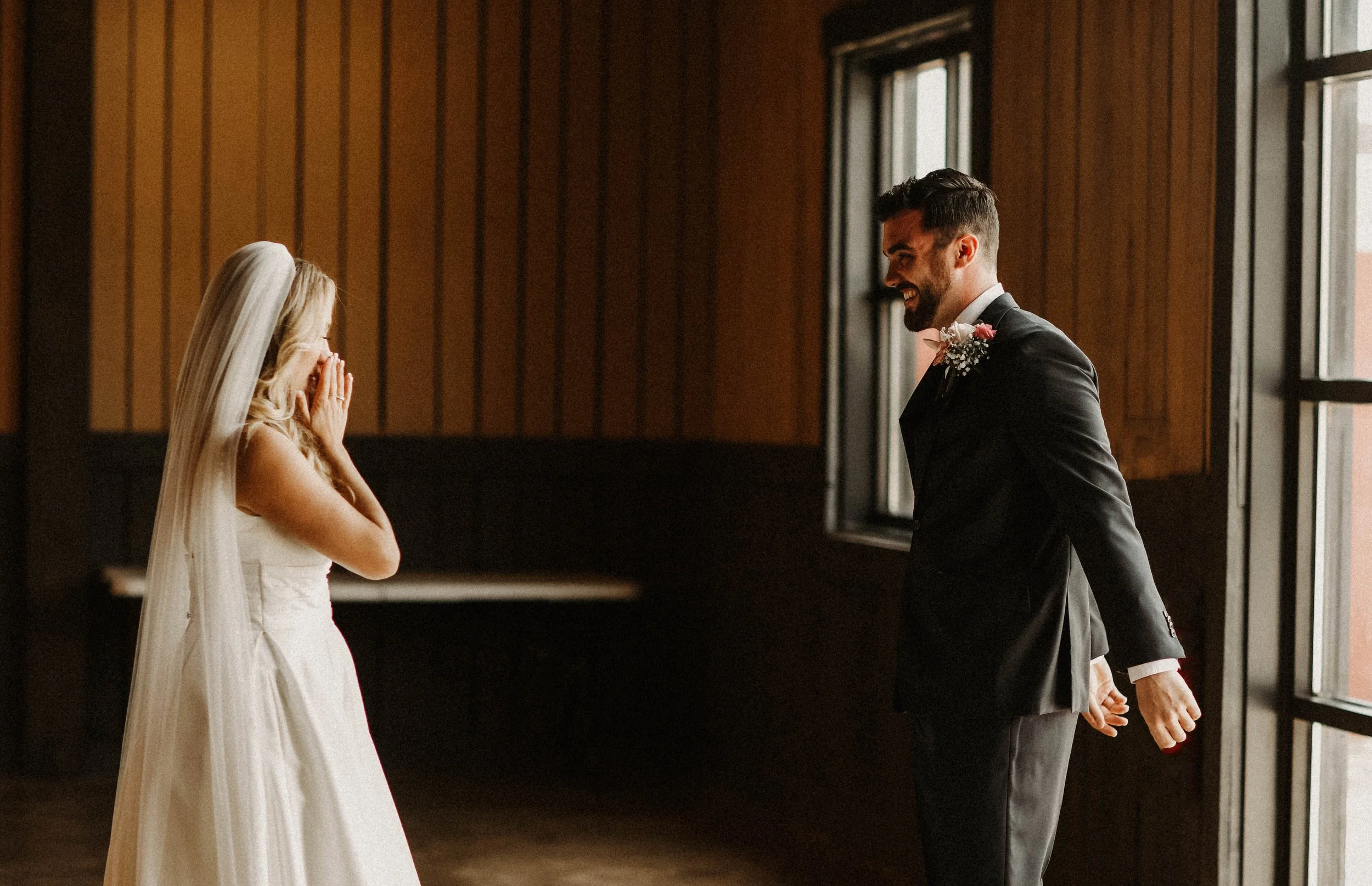 A bride and groom smiling at each other during their wedding ceremony indoors near large windows.