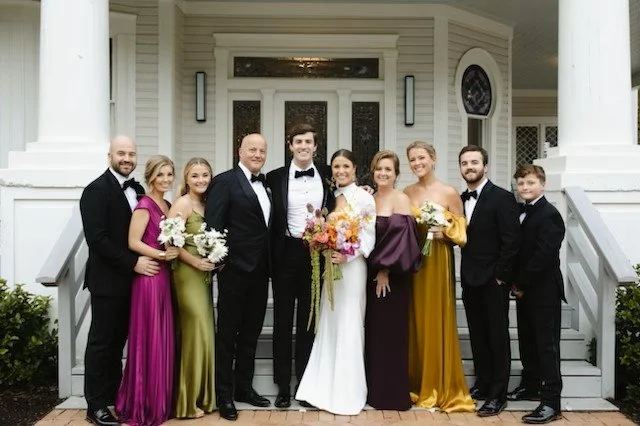 A wedding party with eight people standing in front of a white house. The bride and groom are in the center, surrounded by five women in colorful dresses and two men in black suits. Everyone is smiling.