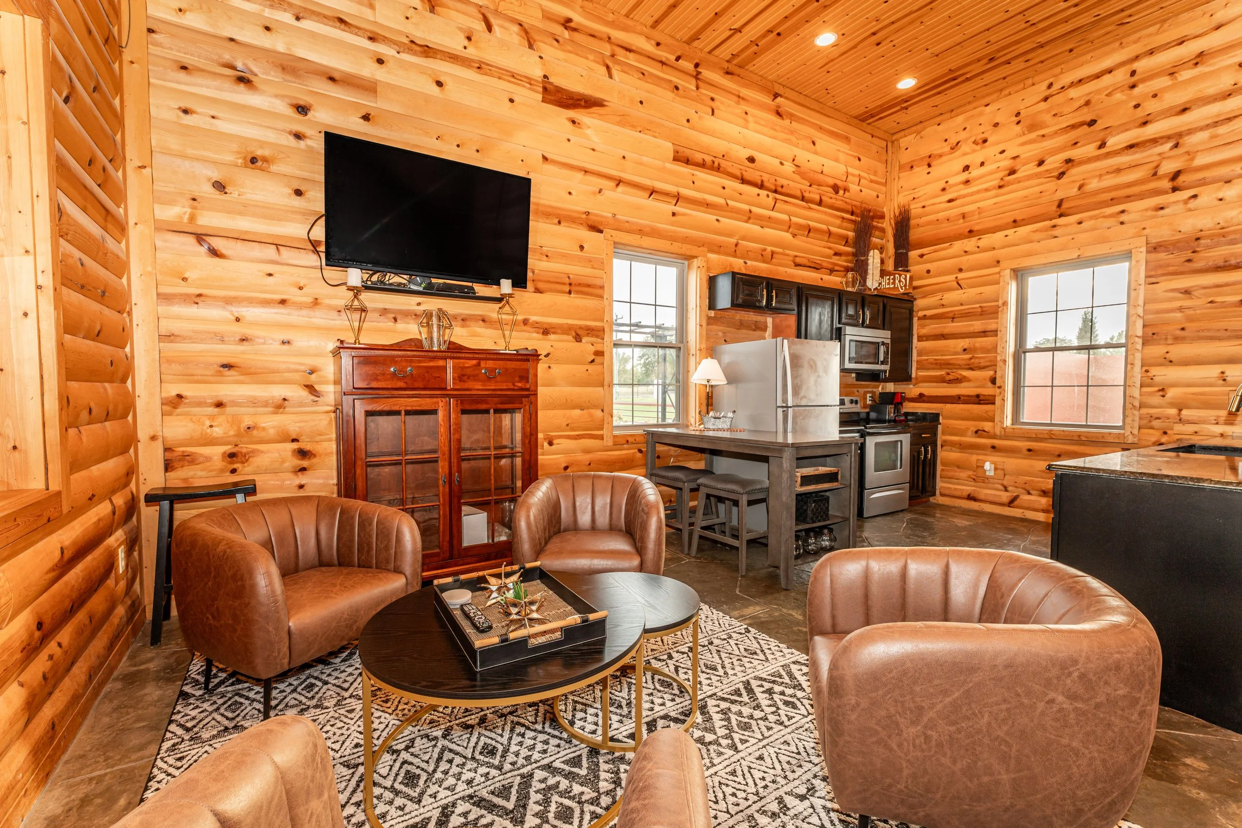 Living room with wooden walls and ceiling, two brown leather armchairs around a black coffee table on a patterned rug, a wooden cabinet, a wall-mounted TV, a window beside the TV, a small dining table with chairs, a kitchen area with a white refrigerator, black cabinets, a microwave, a window, and a decorative sign that says 'Cheers!' with tall dried plants.