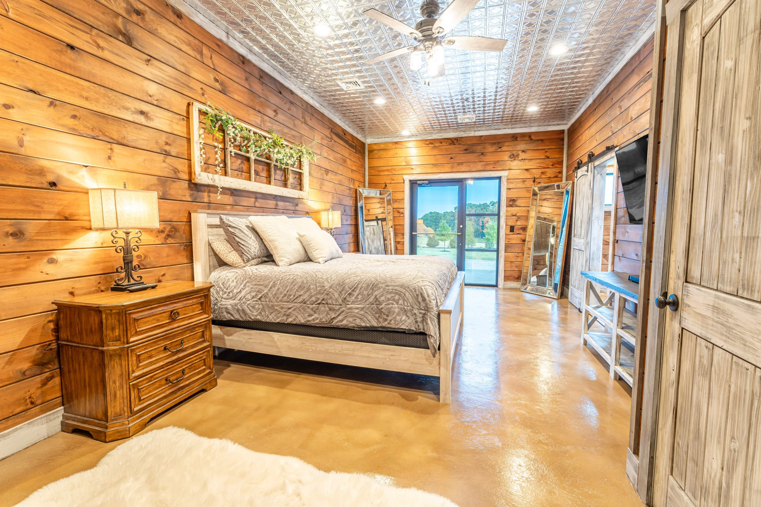 Bedroom with wooden walls, a bed with beige bedding, wooden nightstand with a lamp, large window showing trees outside, full-length mirror leaning against the wall, ceiling fan, and sliding glass door leading outside.