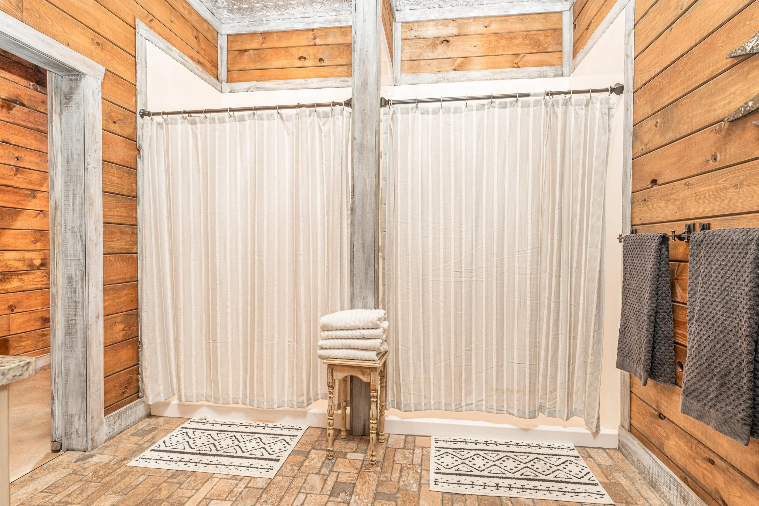 A rustic bathroom with wood-paneled walls and a shower area enclosed by a curtain. There are towels hanging on hooks and a stool with folded towels on top. The floor is made of brick tiles, and there are two small rugs on the floor.