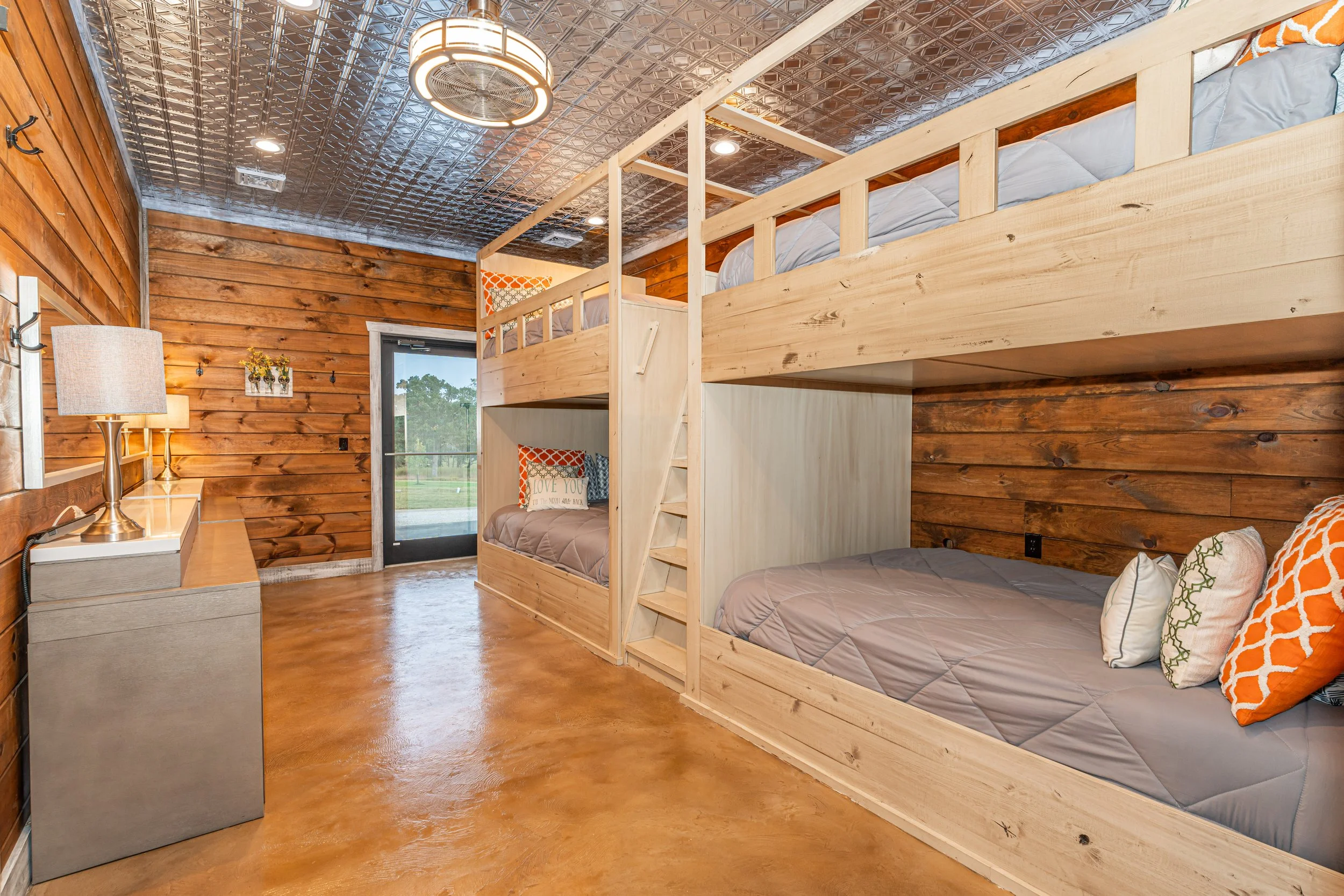 Room with wood-paneled walls and ceiling, featuring a bunk bed with a ladder, single beds with orange and patterned pillows, a window showing trees outside, and a light fixture on the ceiling.