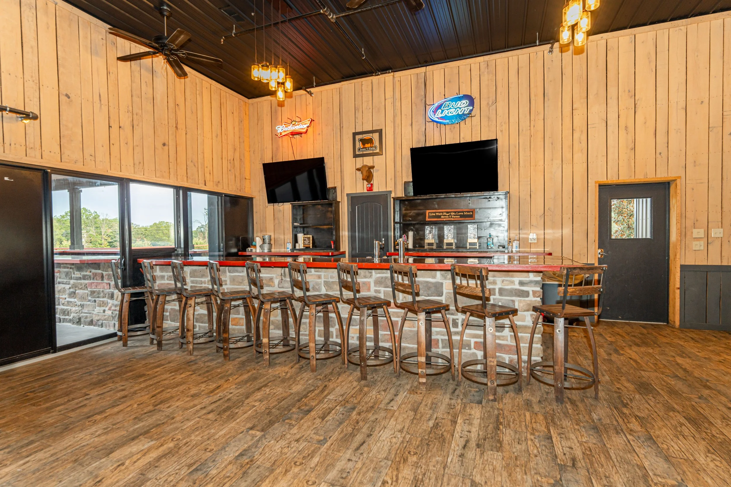 Interior of a bar or restaurant with a long wooden counter, bar stools, two mounted TVs, neon signs including Bud Light and Budweiser, a cow head wall decor, and large windows with outdoor greenery visible.