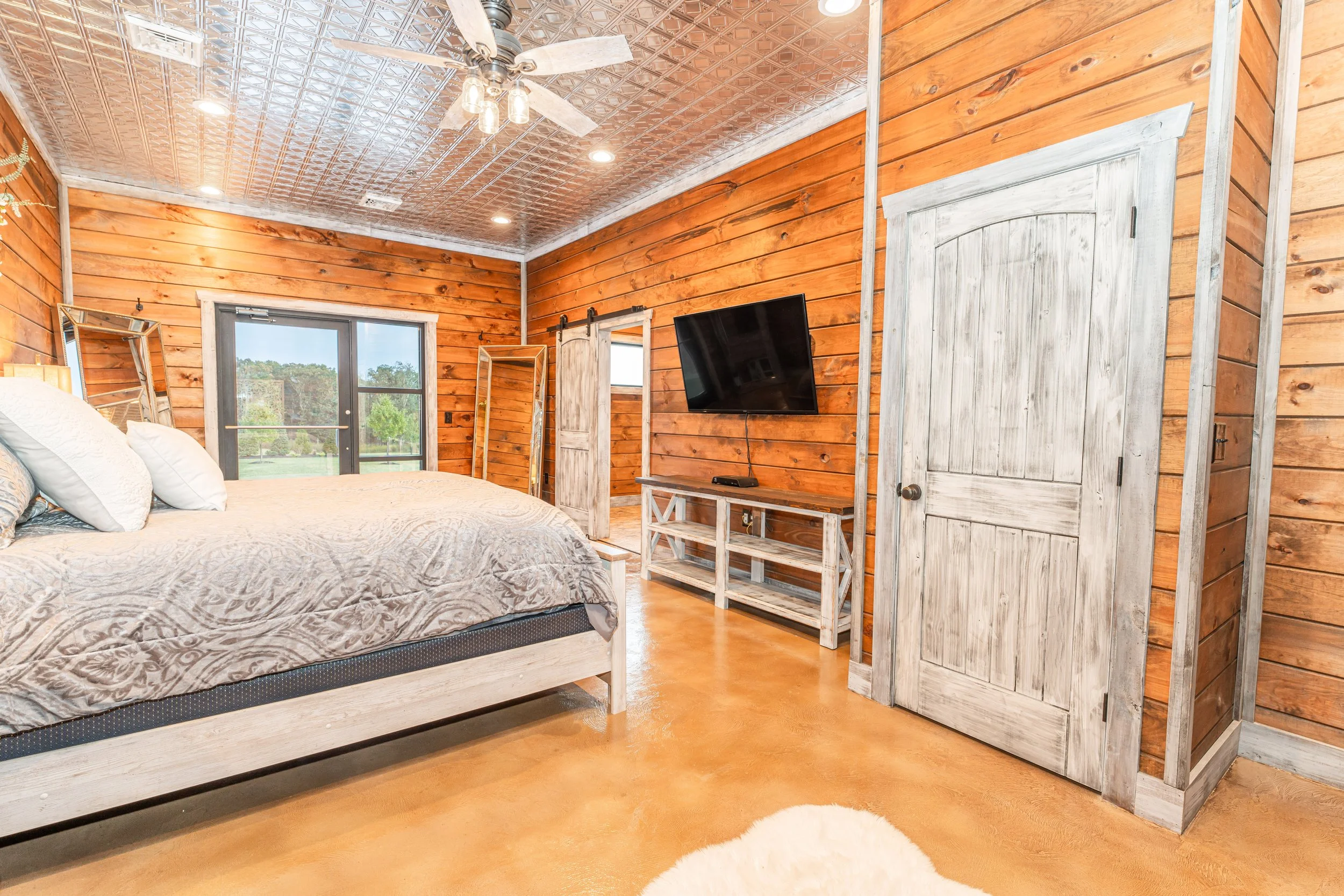 A cozy bedroom with wood-paneled walls and ceiling, a bed with decorative pillows, a sliding glass door leading outside, a wall-mounted flat-screen TV, a rustic wooden door, and a fluffy white rug on the wooden floor.