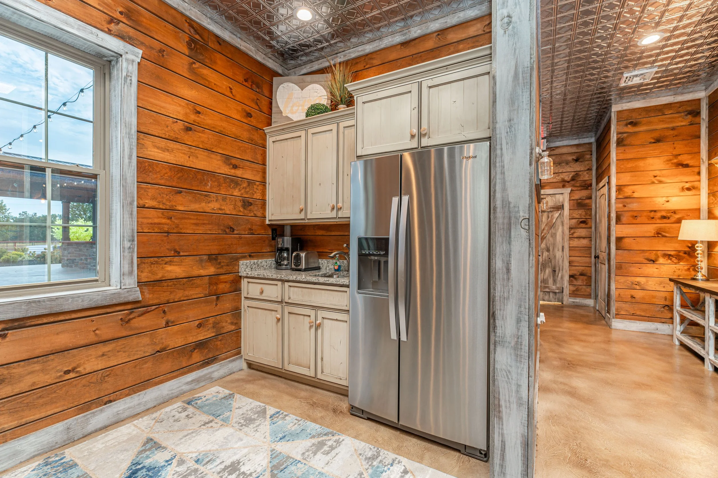 Interior view of a kitchen with wood-paneled walls, a stainless steel refrigerator, white cabinets, a window, and a geometric patterned ceiling.